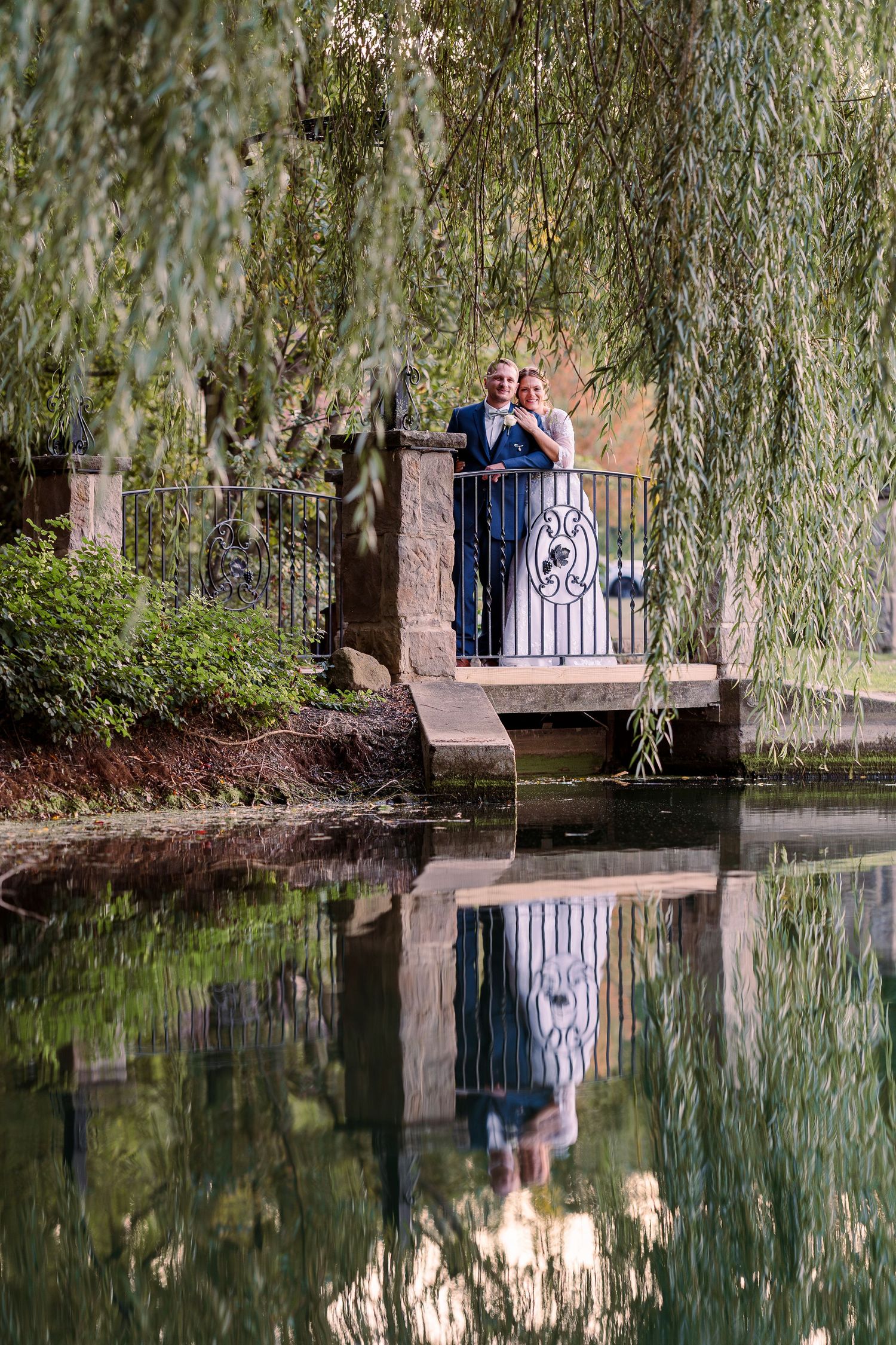 Bride and groom standing on a stone bridge over a pond framed by weeping willow trees at Gervasi Vineyard wedding venue