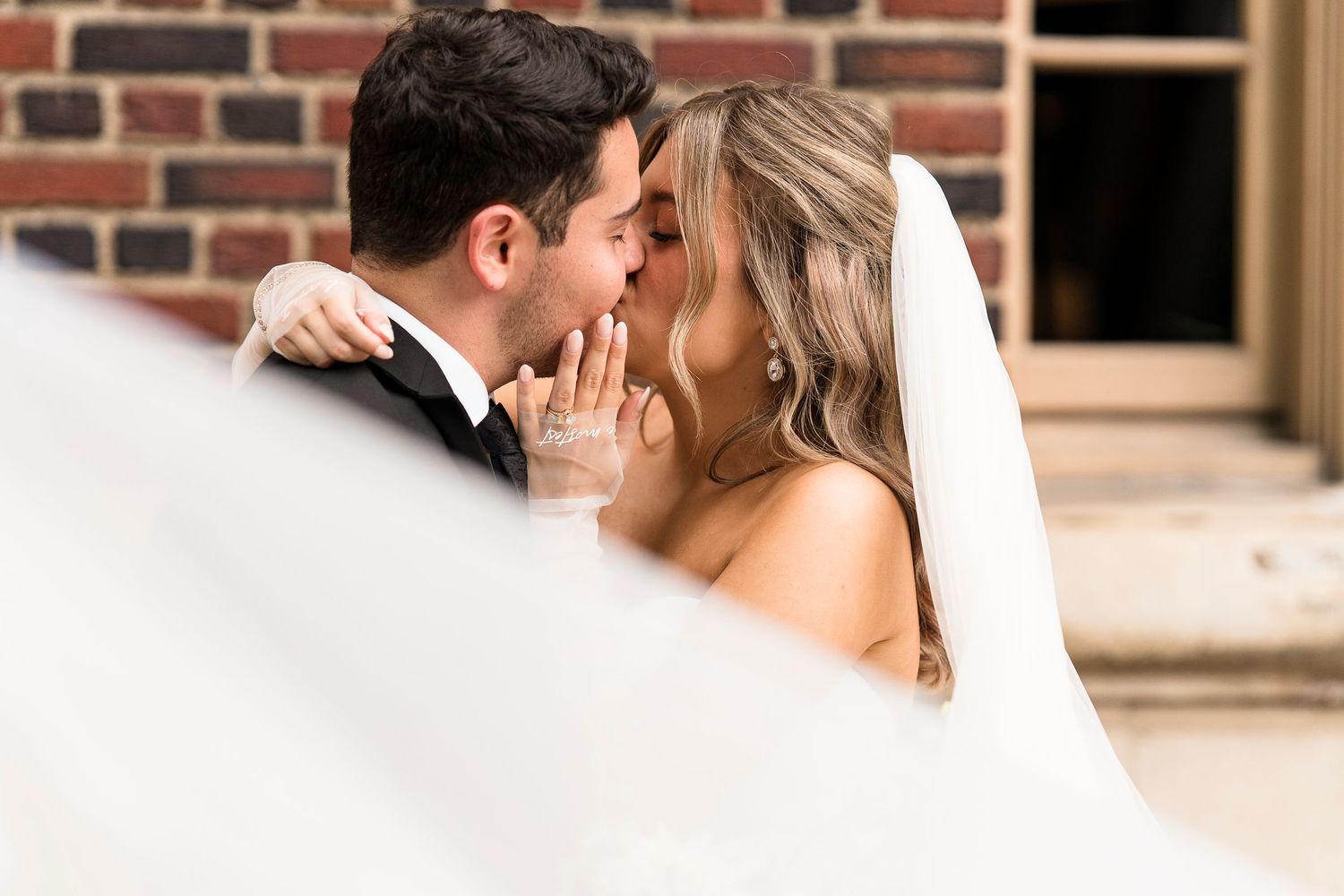 Bride and groom sharing a romantic kiss, bride wearing veil, against a brick wall backdrop.