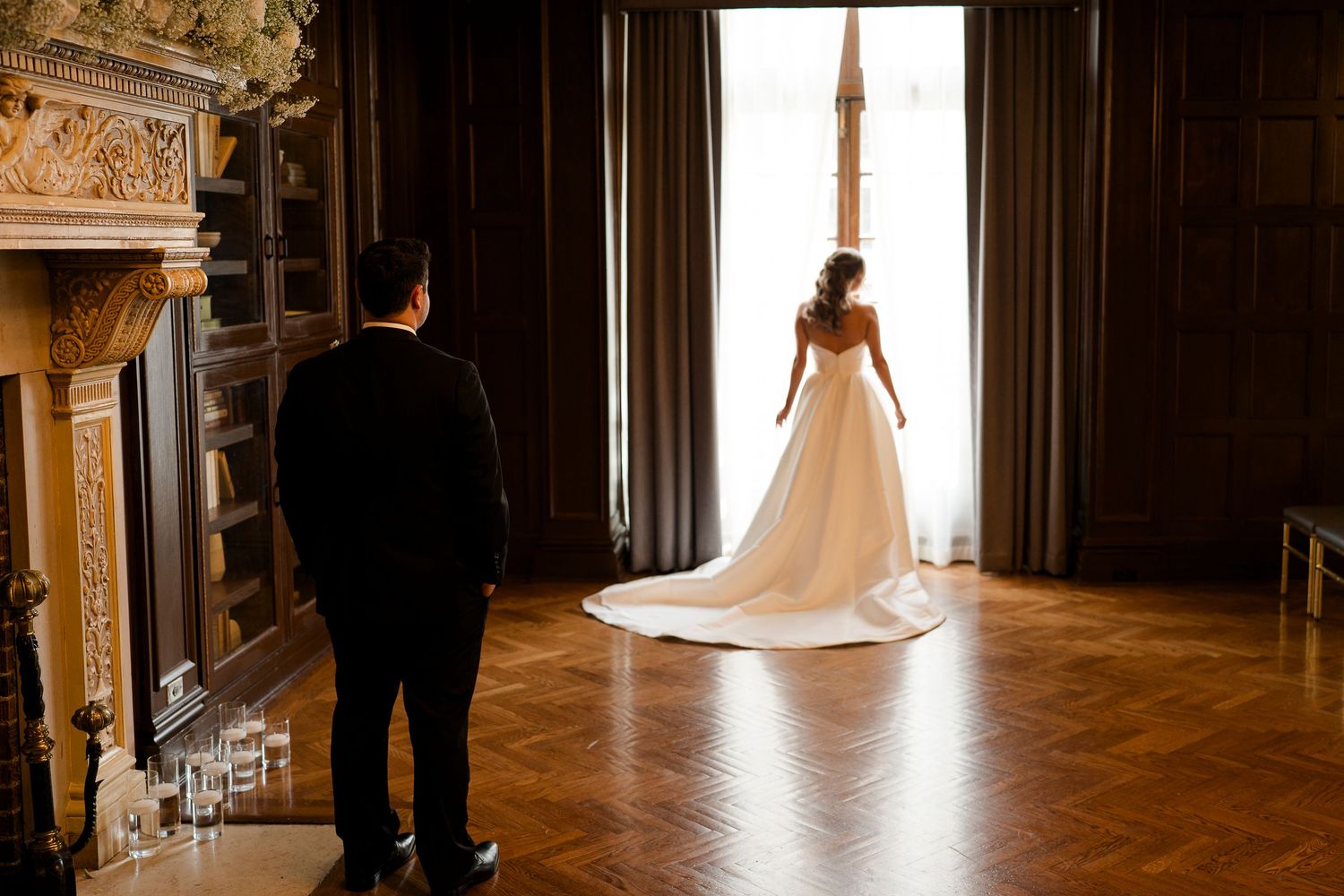 Bride in elegant white gown stands by grand window as groom looks on in ornate ballroom.