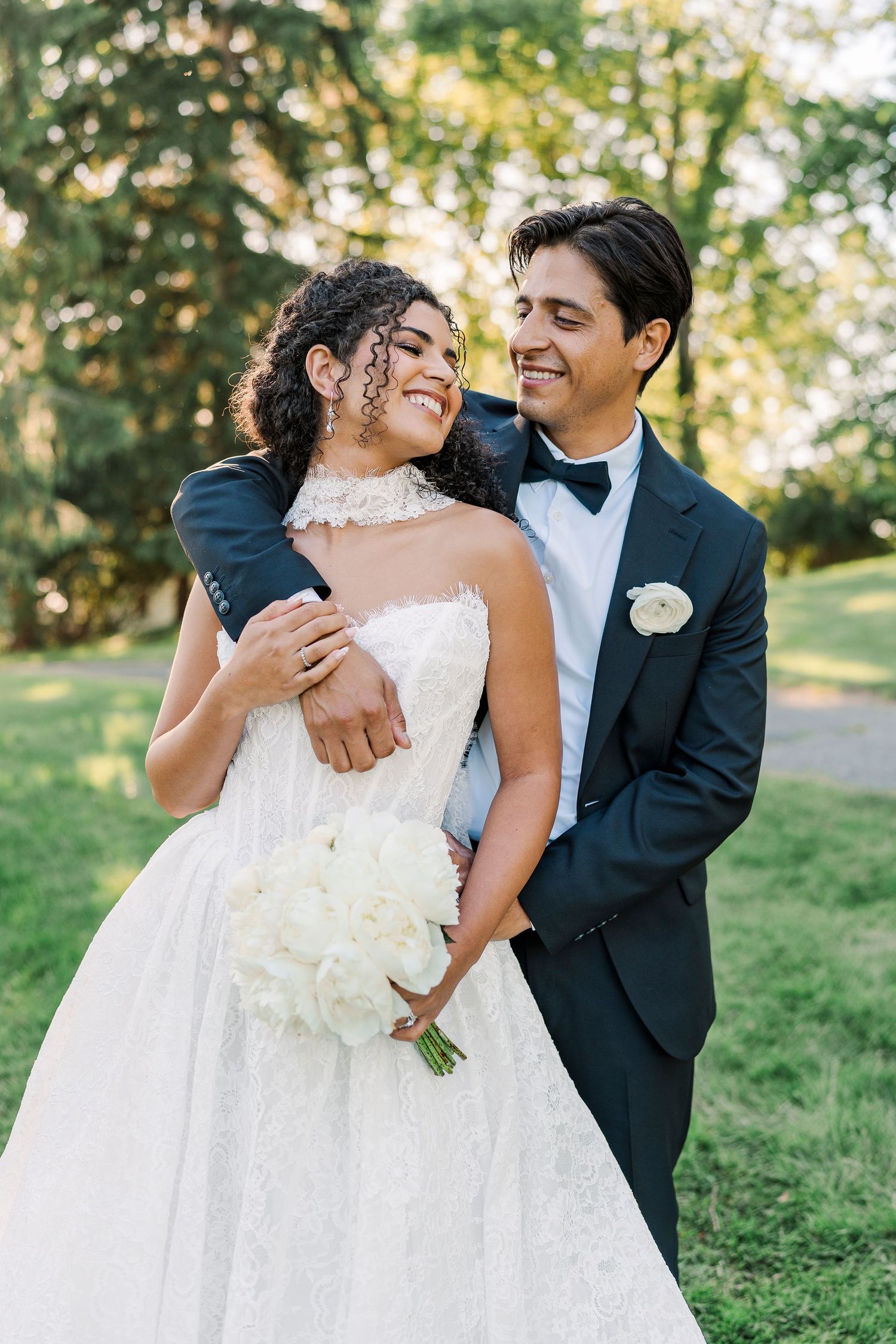 A smiling bride in a white lace gown holds a bouquet while a groom in a navy tuxedo embraces her outdoors.