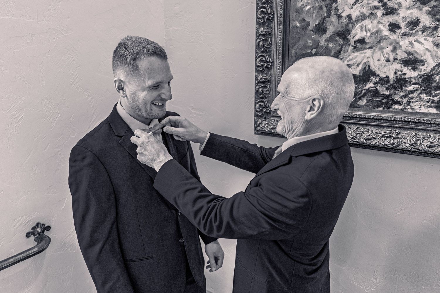 Black and white photo of older man adjusting groom's bow tie before wedding.