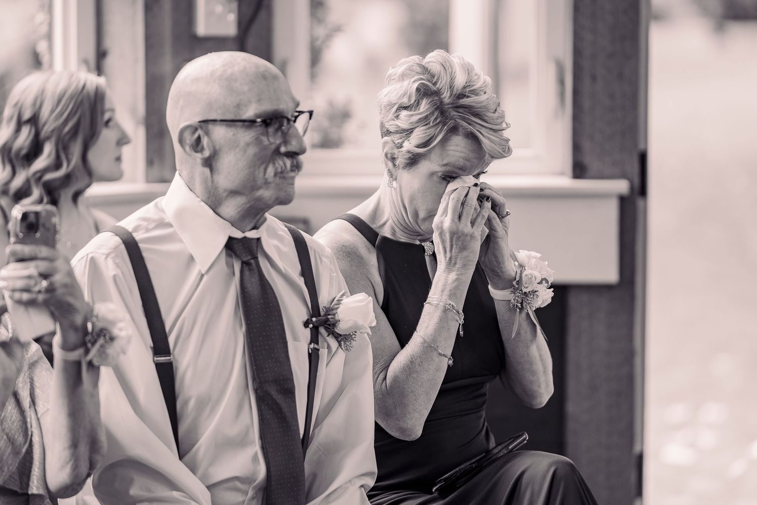 Emotional wedding guests, man in suspenders and woman dabbing tears at ceremony.