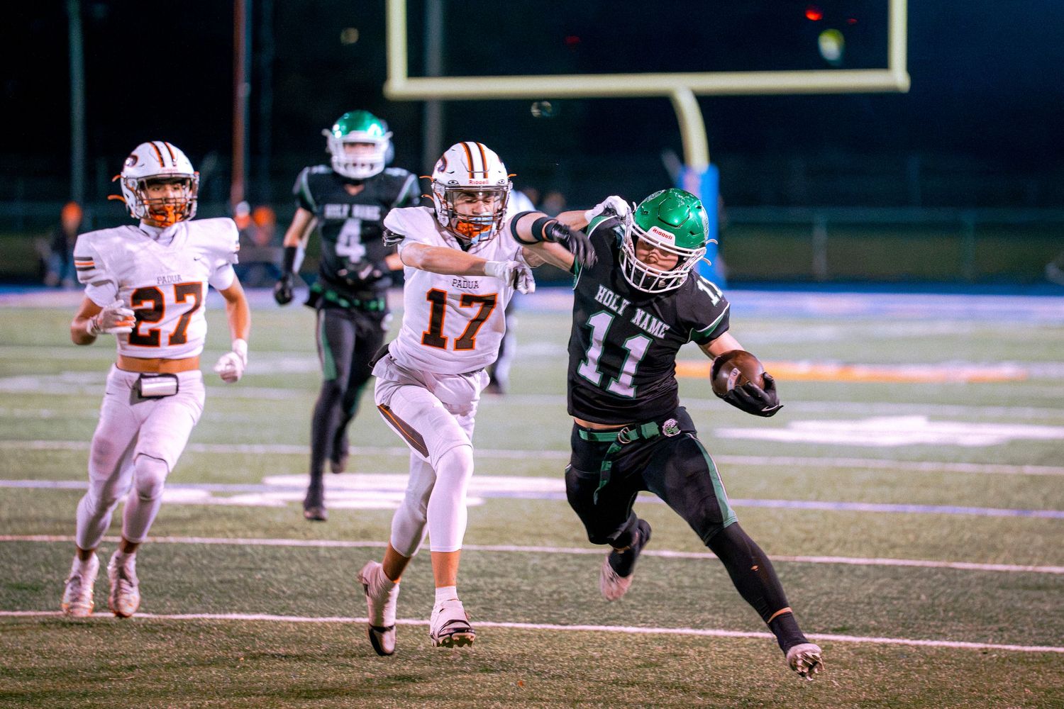 A Holy Name football player in green jersey number 11 runs past Padua defenders during a night game under stadium lights.