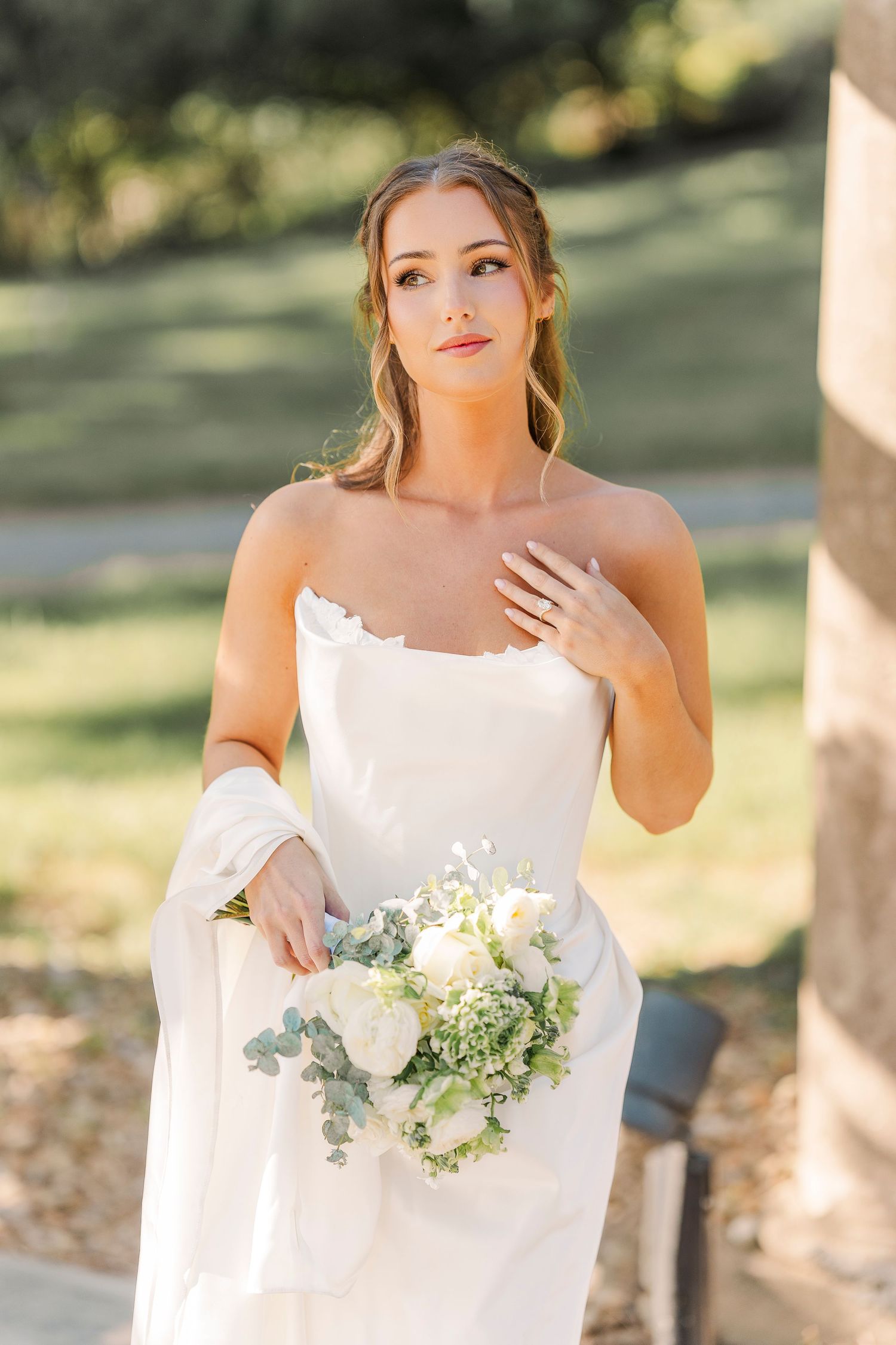 Elegant bride in strapless white gown holds white floral bouquet outdoors in soft sunlight.