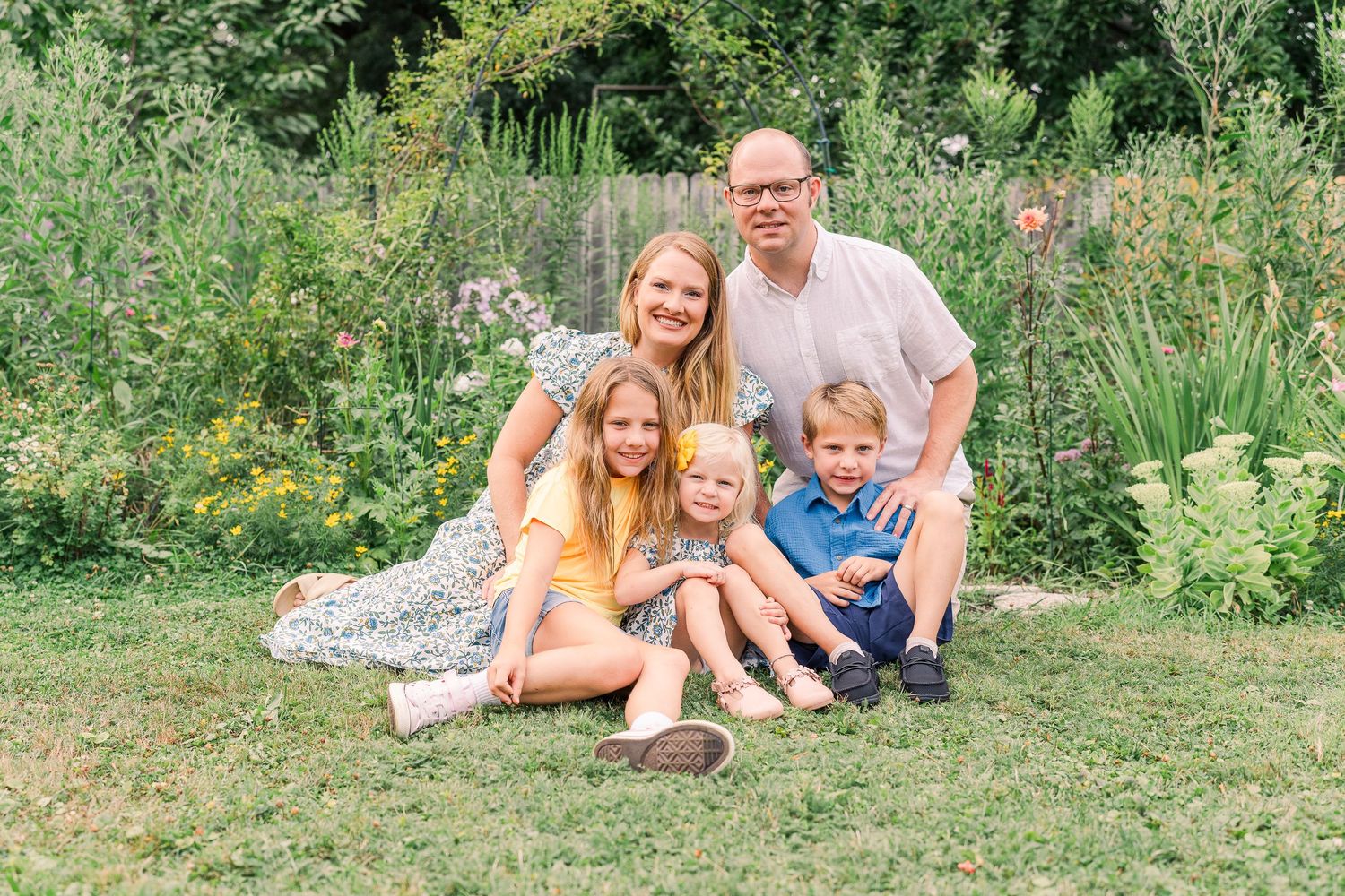 Smiling family of five posing together on green lawn with lush flower garden backdrop in summer.