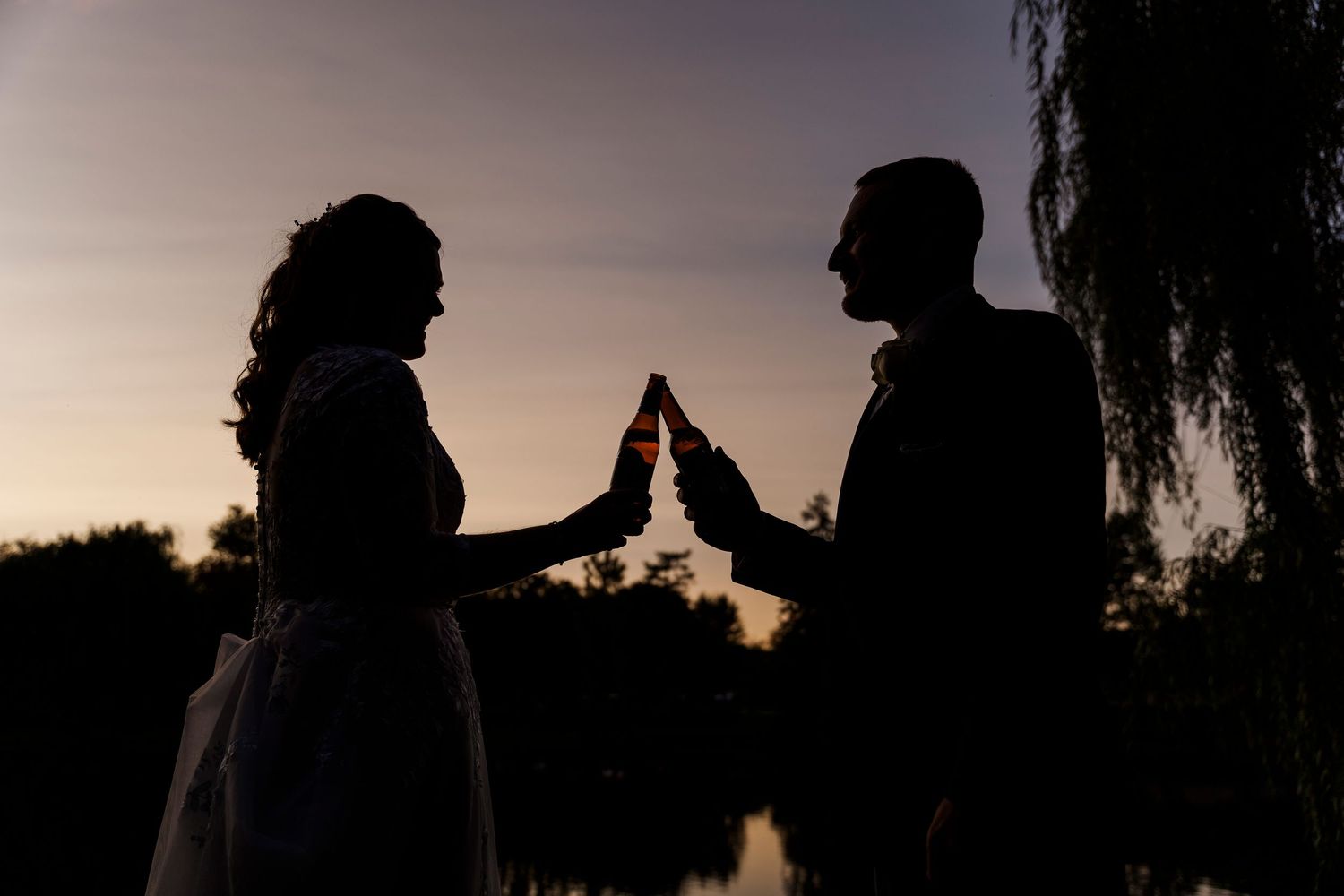 Silhouette of bride and groom toasting at sunset beside a serene reflective pond.