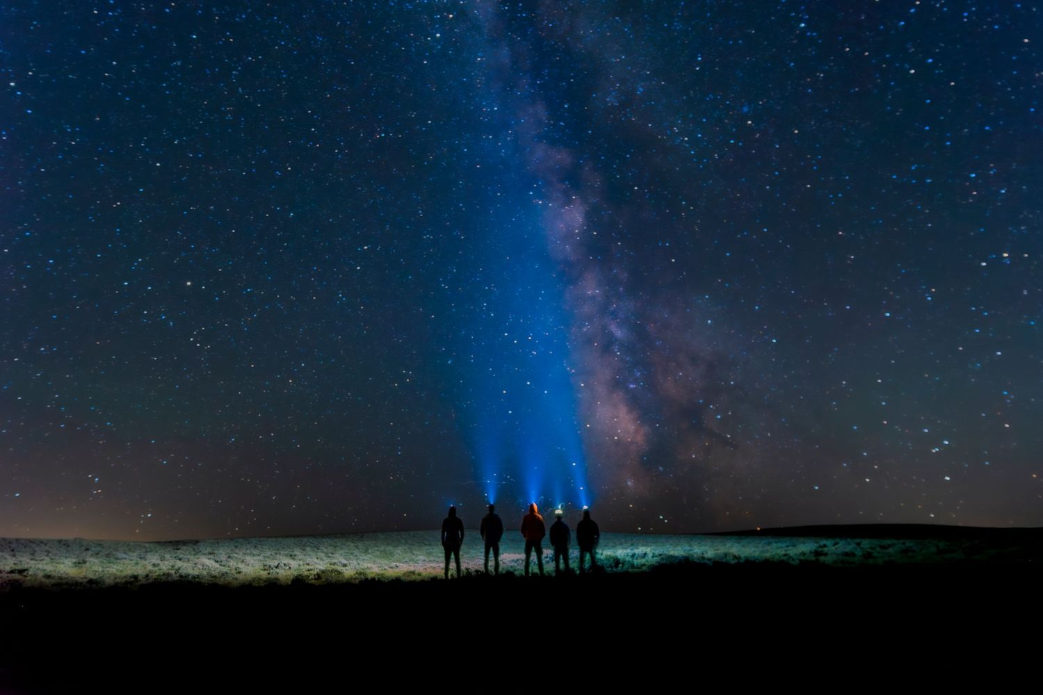 Five silhouetted figures stand under a stunning Milky Way night sky, shining flashlights toward glowing stars.