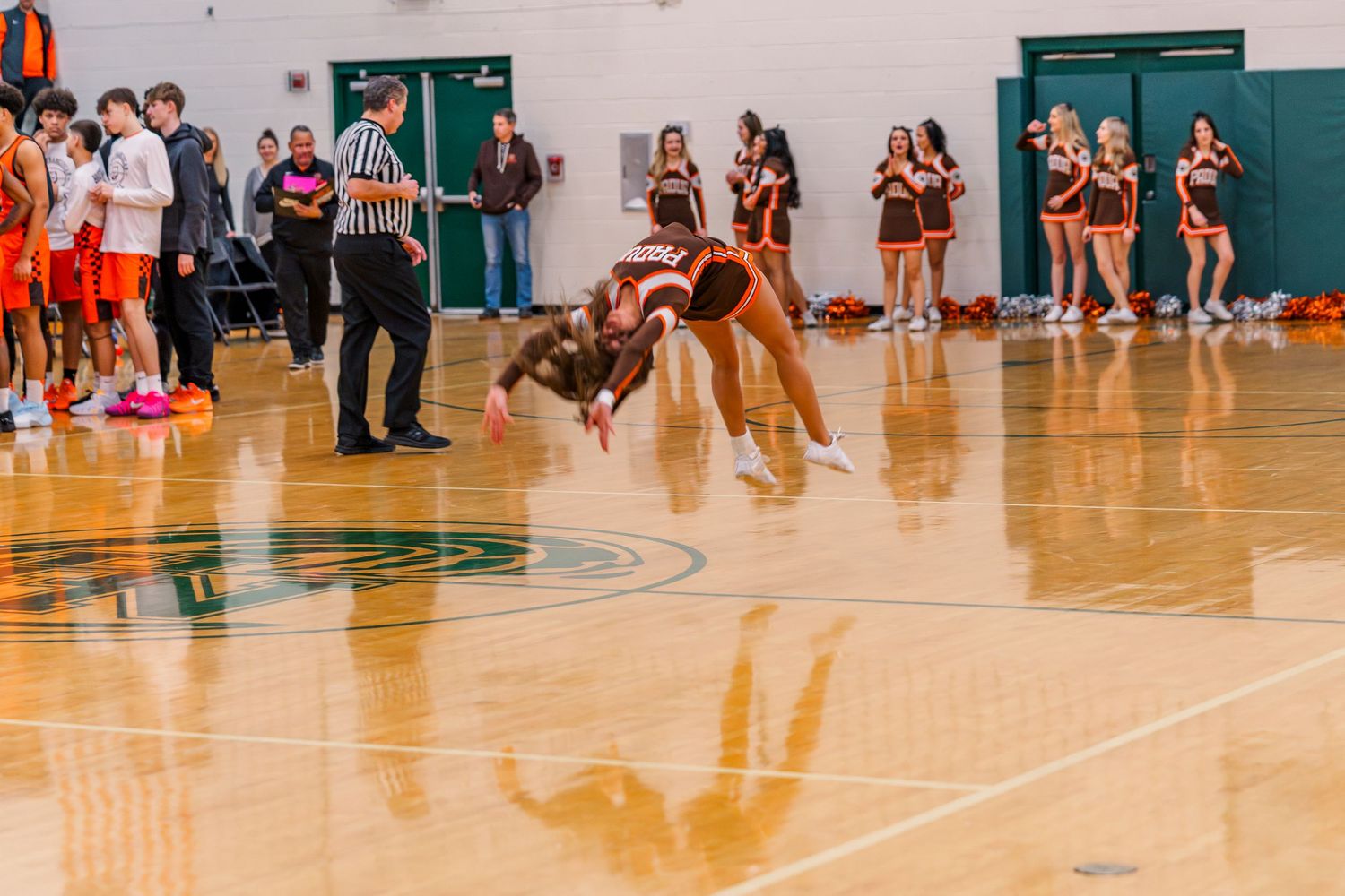 Padua Cheerleaders perform an acrobatic stunt on a green gym floor during a basketball game with teammates cheering in background.