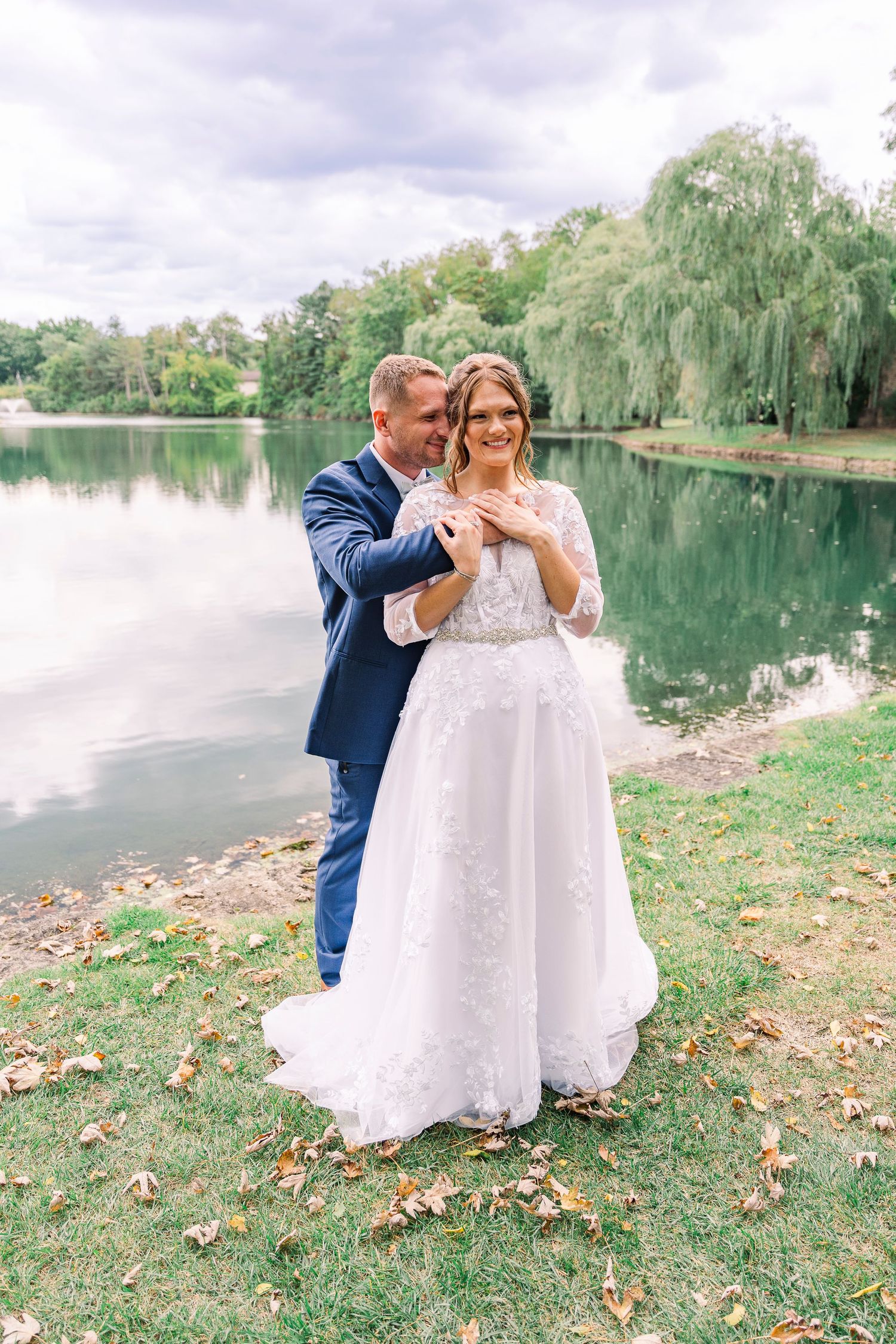 Happy bride and groom embracing by a serene lake surrounded by lush green trees.