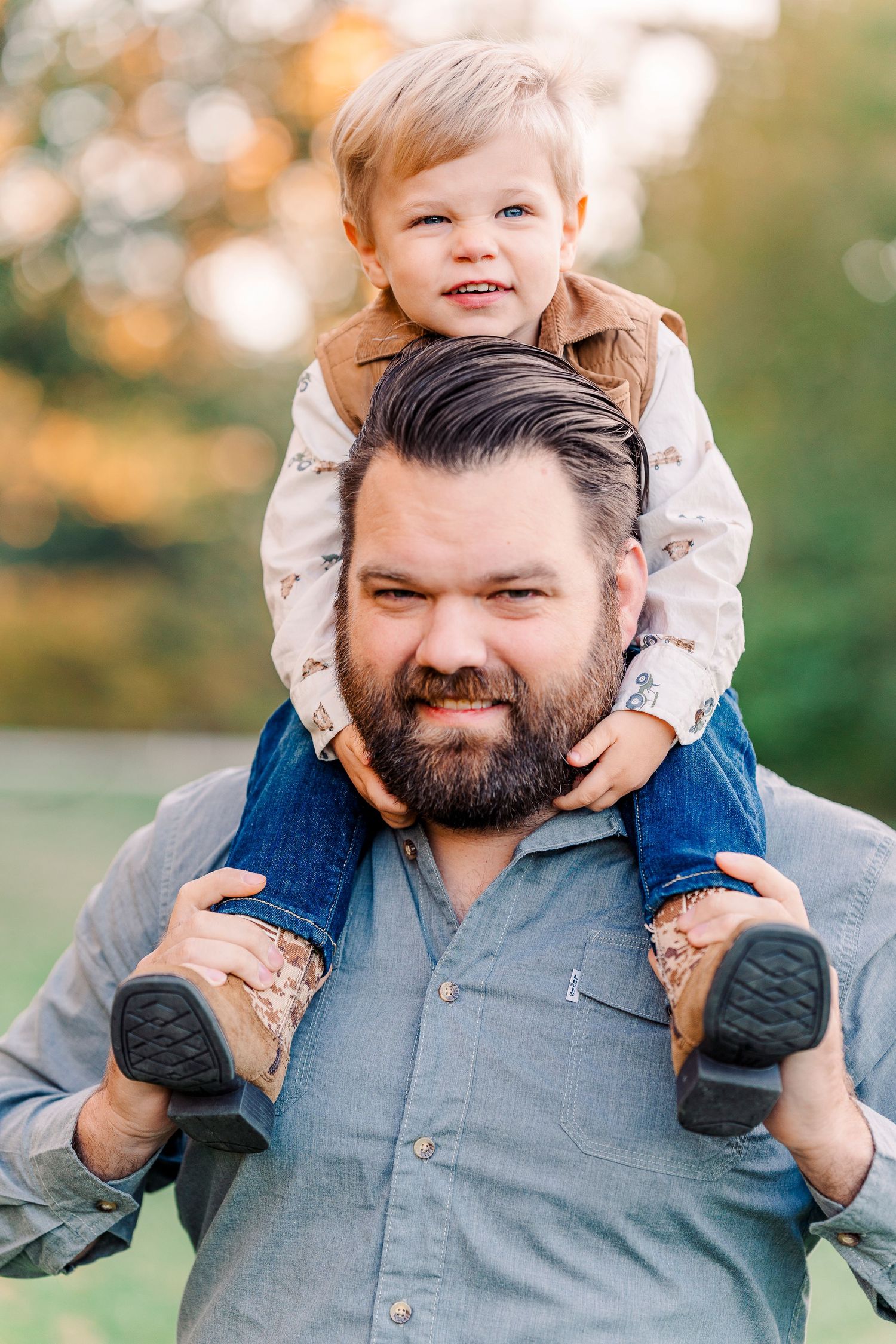 A bearded man carries a happy toddler on his shoulders outdoors with a blurred green background.