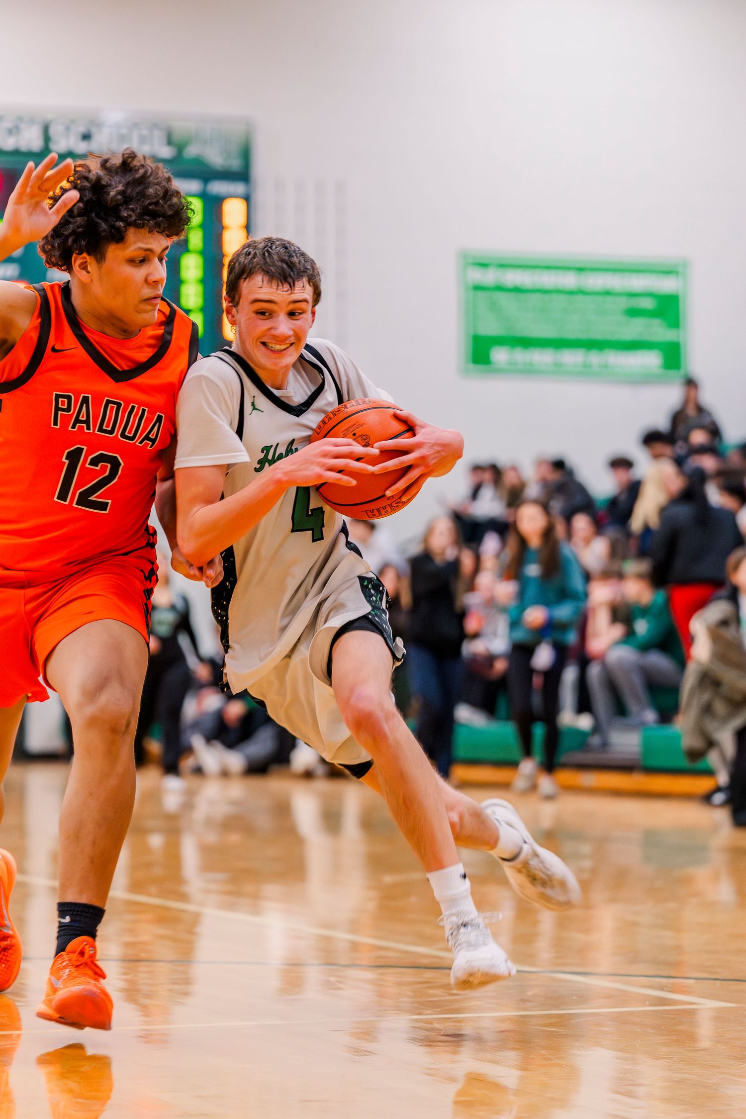 Holy Name High school basketball player in green dribbles past Padua defender wearing orange jersey number 12 during indoor game.