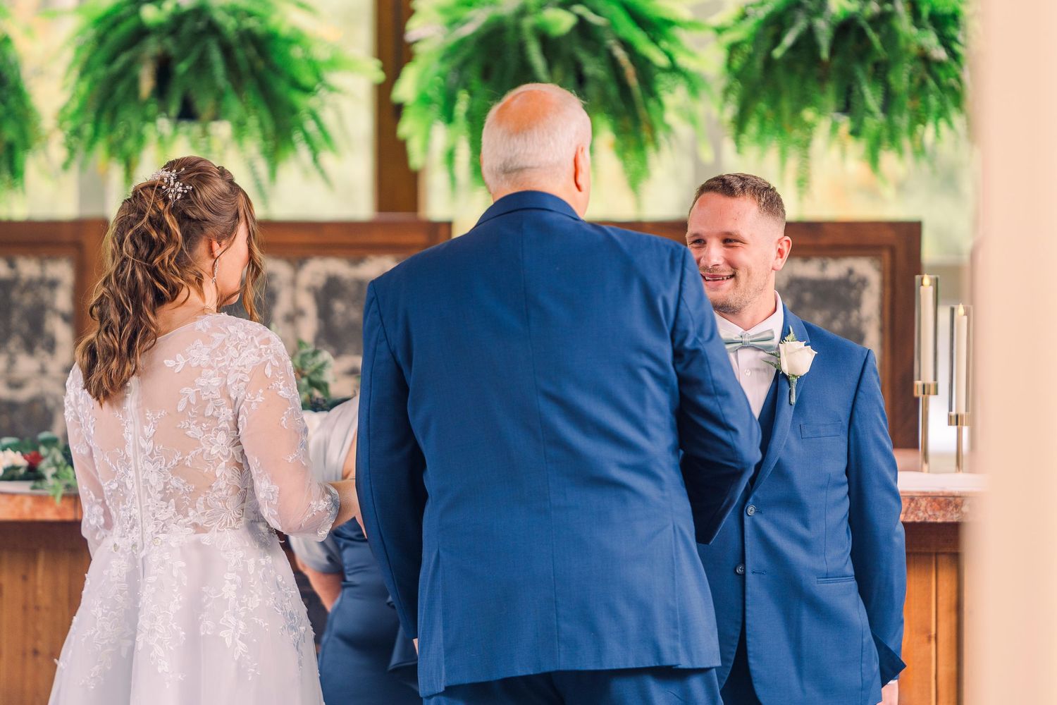Groom in navy suit smiles as bride and escort approach him at the wedding ceremony altar.