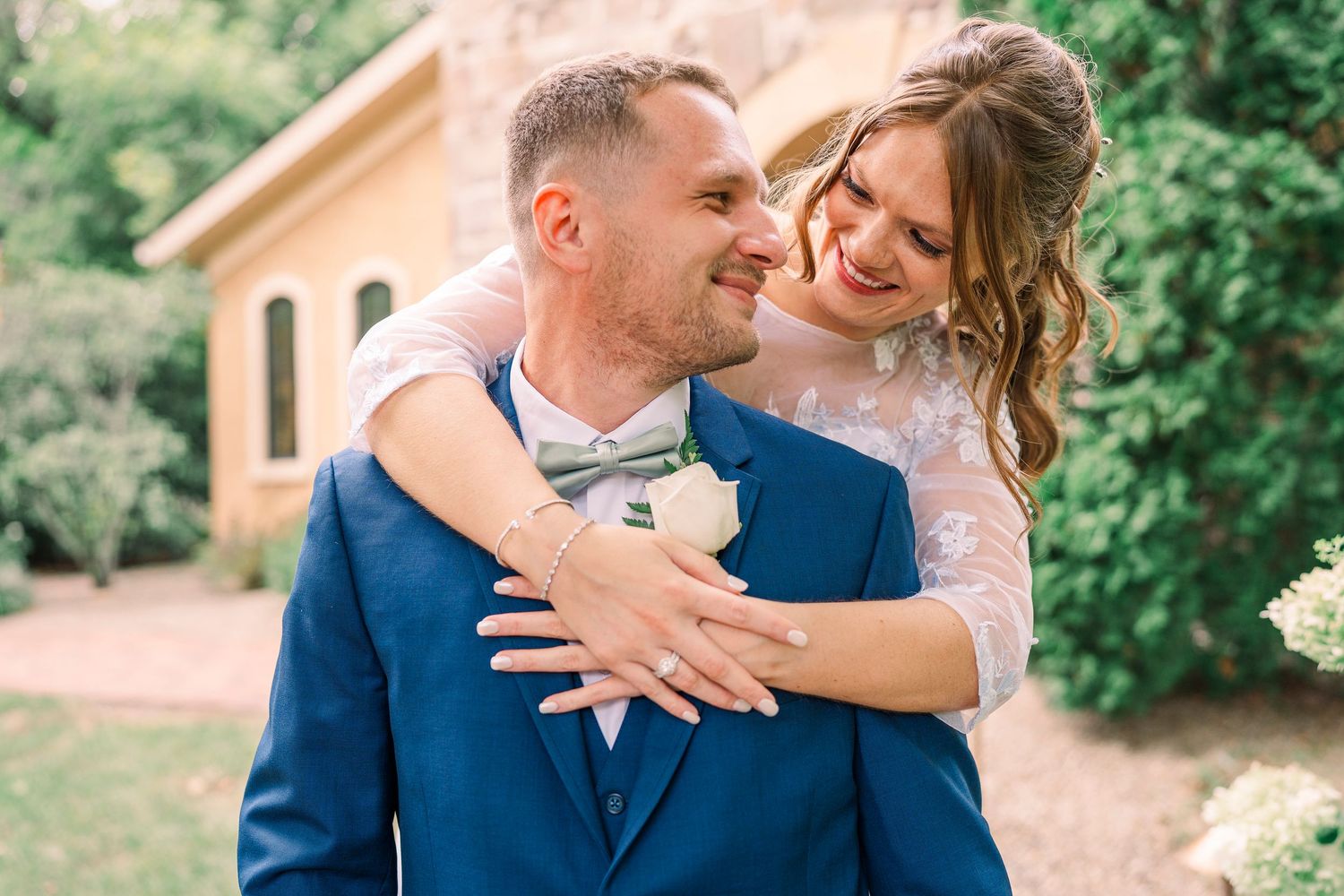 Happy bride embracing groom in blue suit with boutonniere in outdoor garden setting.