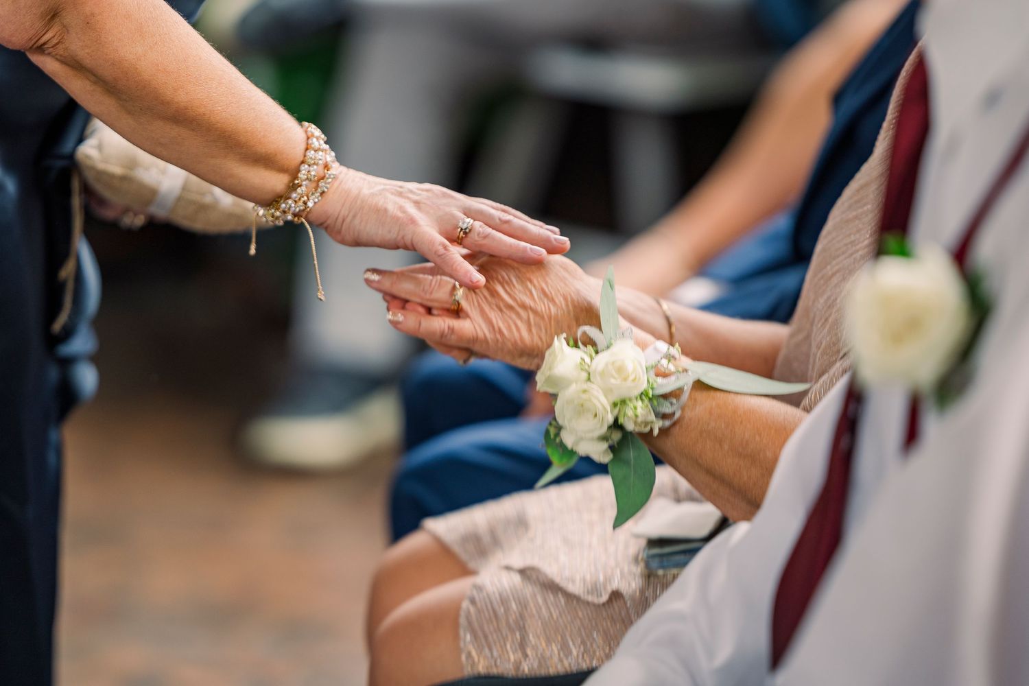 Close-up of wedding guests holding hands with floral wrist corsage at ceremony during warming of the rings ceremony.