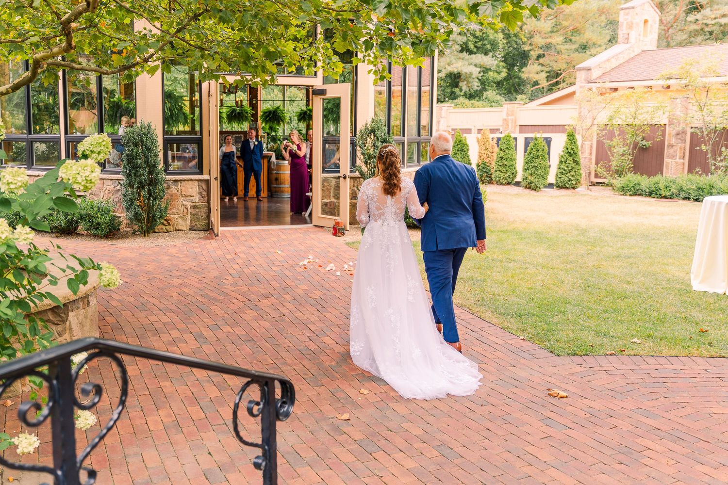 Bride and escort in navy suit walk toward the conservatory ceremony venue at on brick pathway at Gervasi Vineyard.