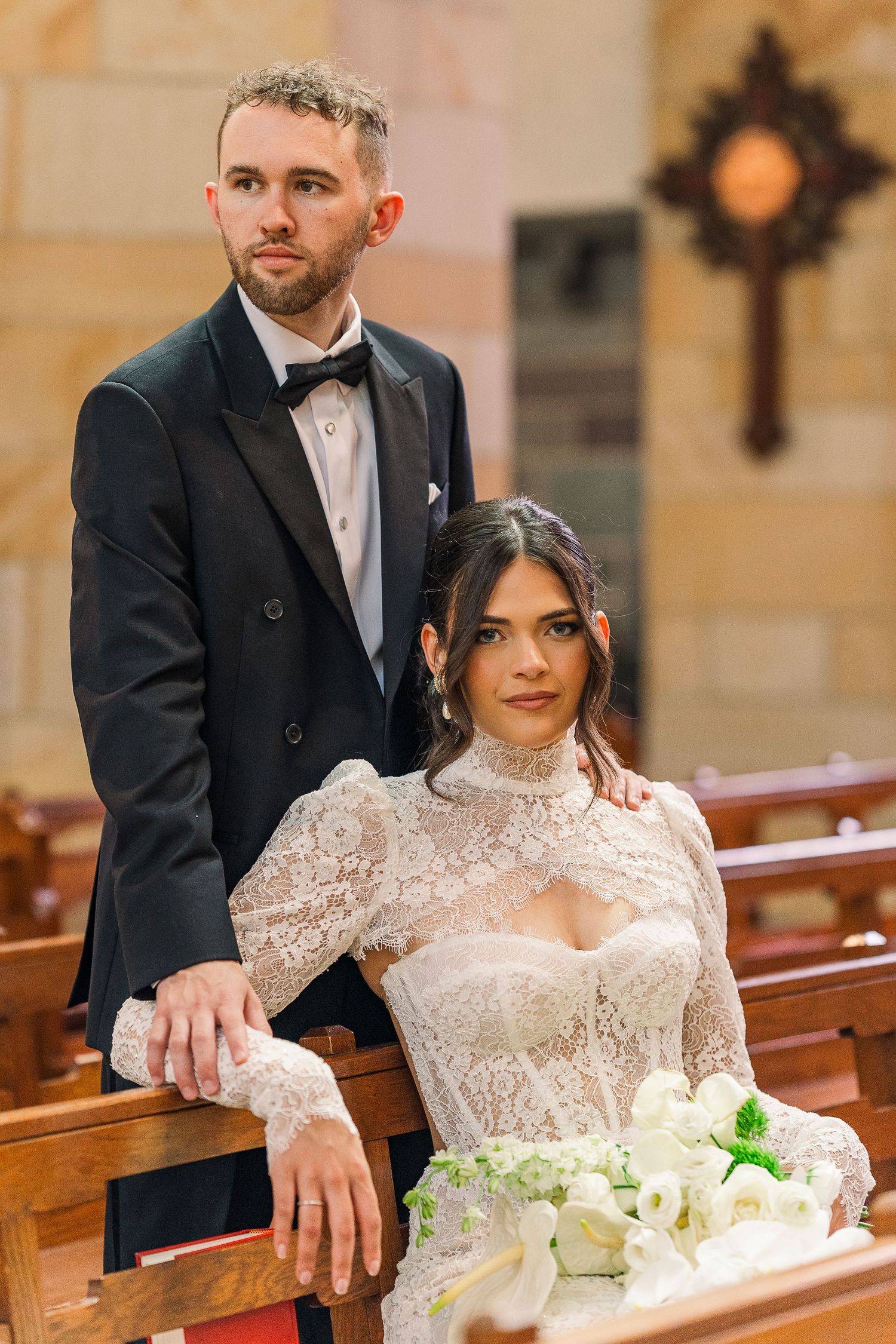 Bride and groom posing elegantly in a church pew, bride wearing a lace high-neck gown holding white flowers.