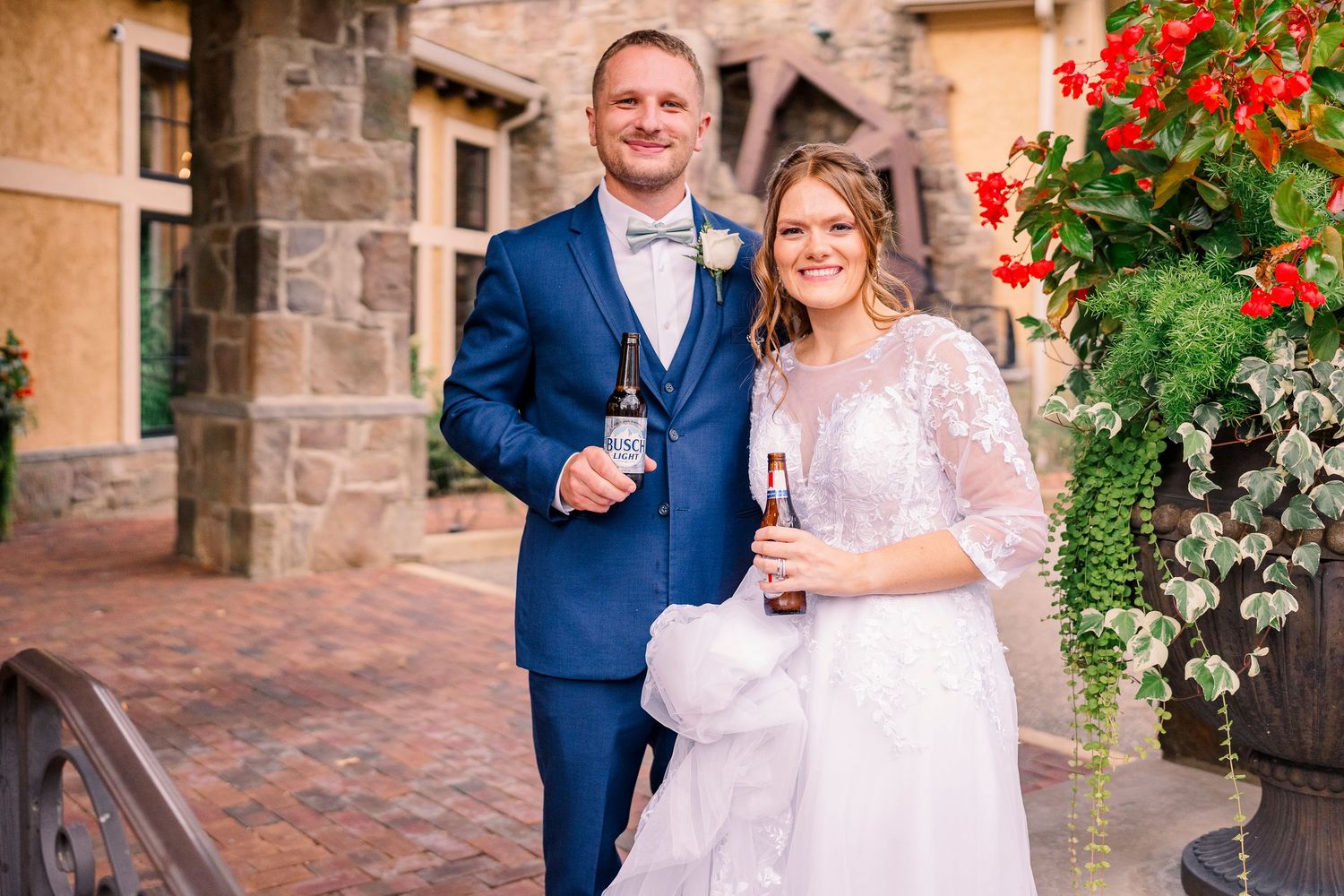 Bride and groom smiling and holding champagne glasses outside a stone building with red flowers.