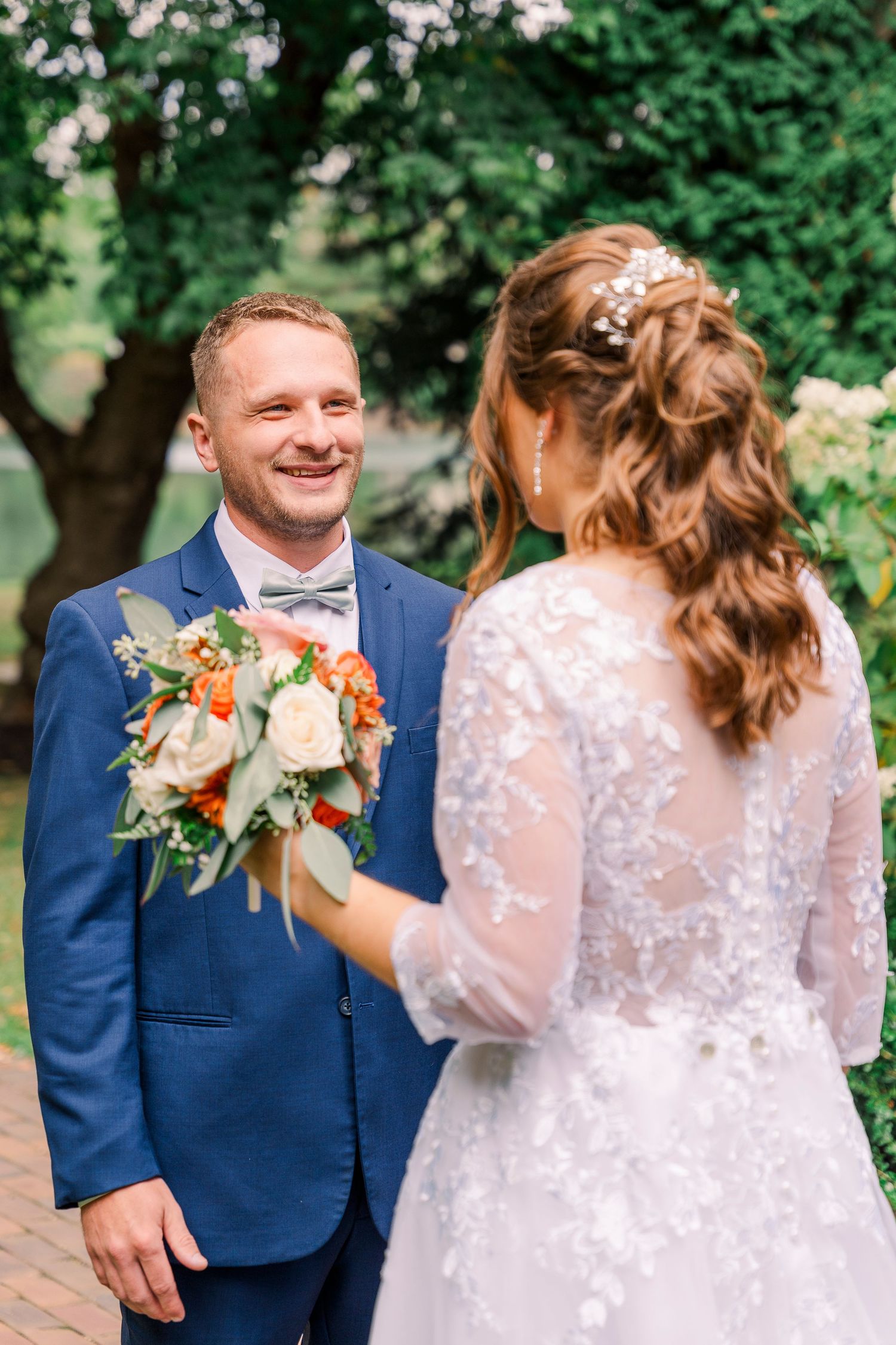 Smiling groom in blue suit holding bouquet facing bride in lace wedding dress.