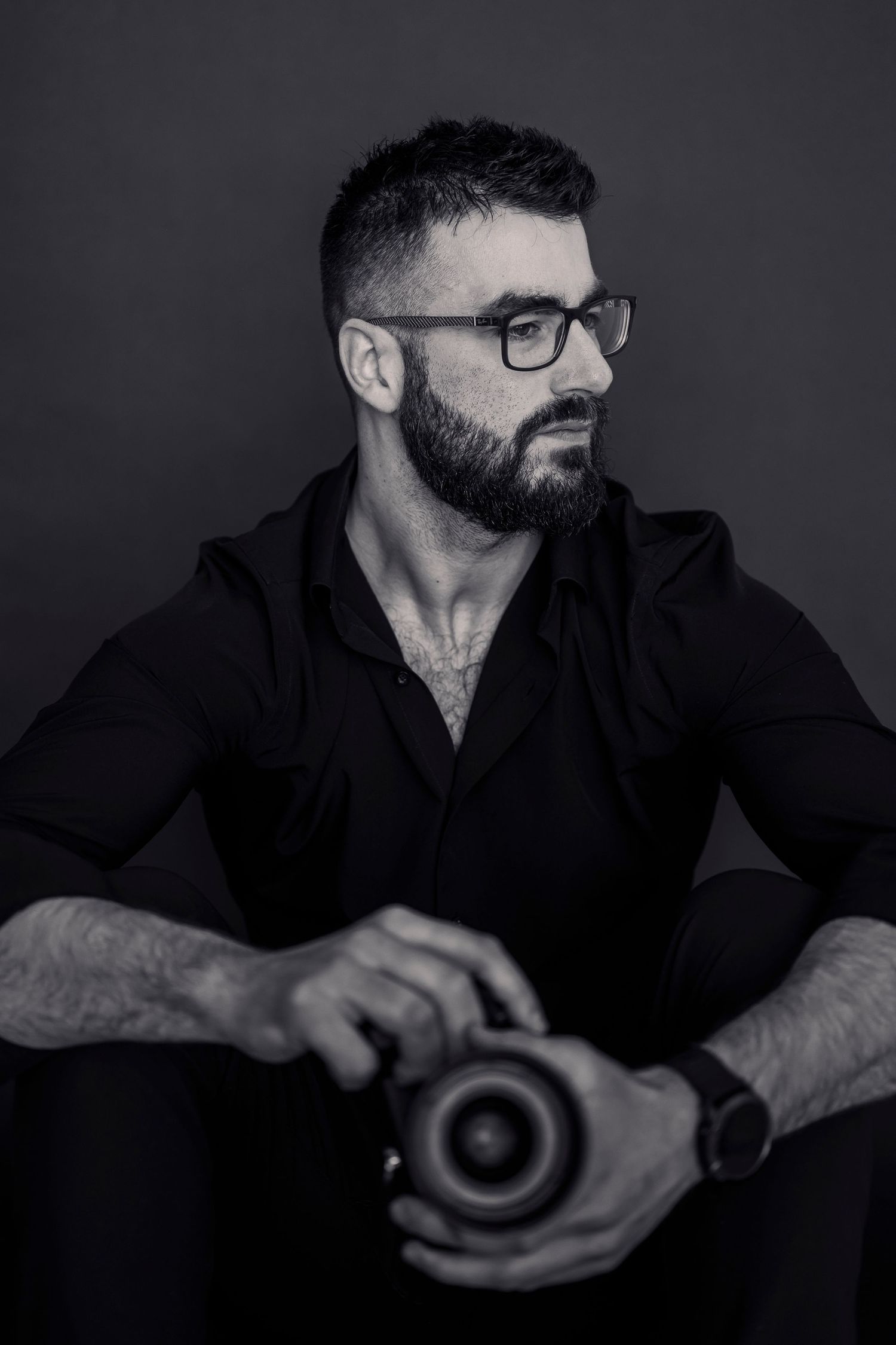 Black and white portrait of bearded man with glasses holding camera lens.