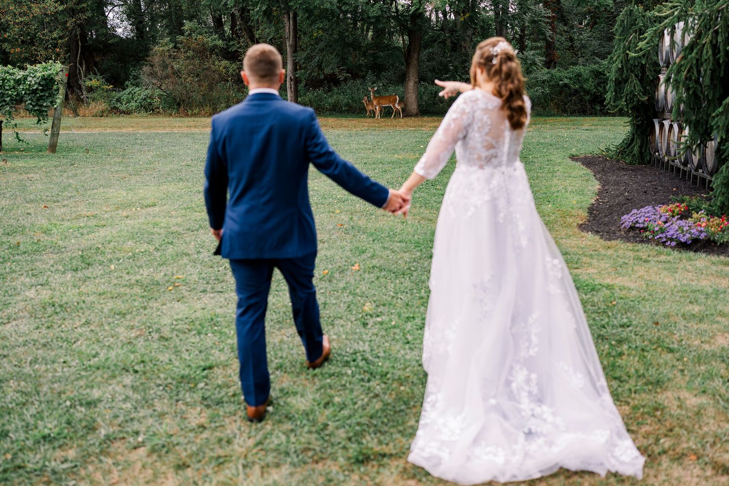 Bride and groom walking hand in hand across a green lawn, bride pointing into the distance.