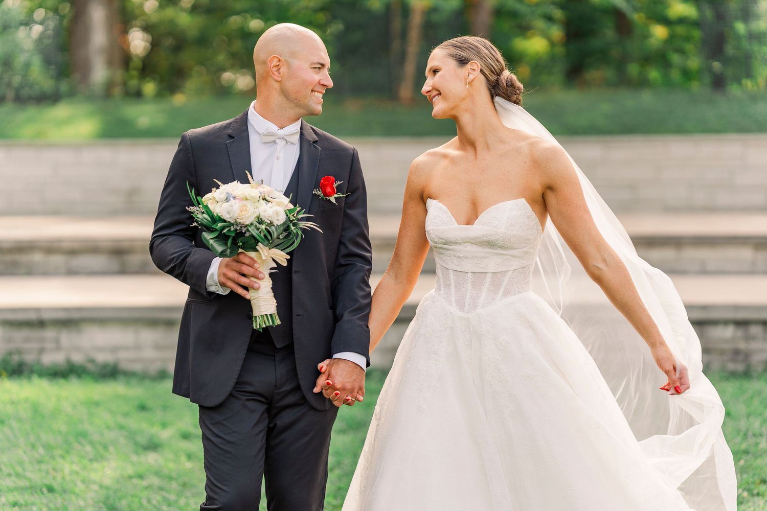 Bride and groom holding hands and bouquet, smiling at each other outdoors on their wedding day.