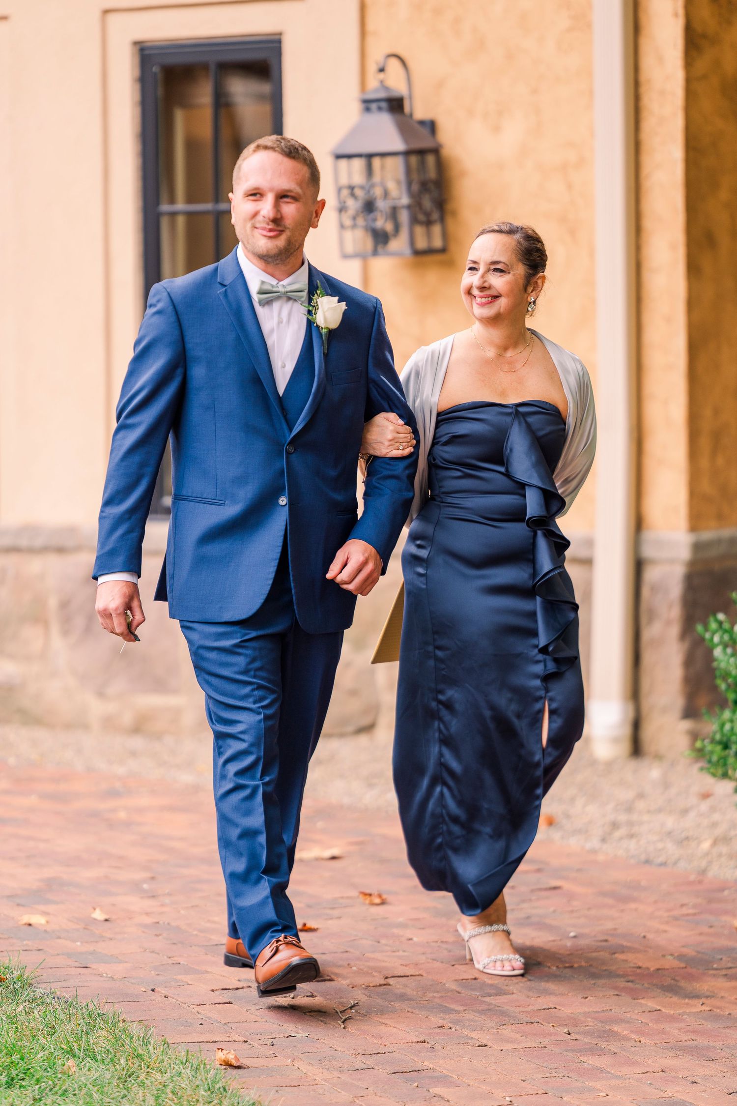 Groom in navy suit with bow tie walks arm-in-arm with woman in navy dress on brick pathway.