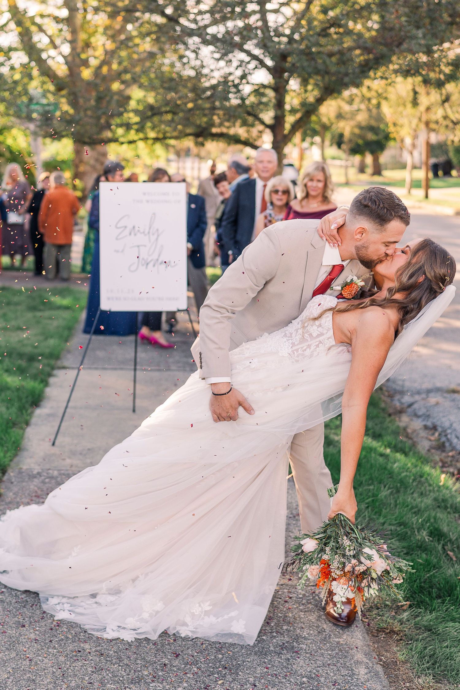 Bride and groom share a romantic dip kiss outdoors after their wedding ceremony, with guests celebrating in the background.