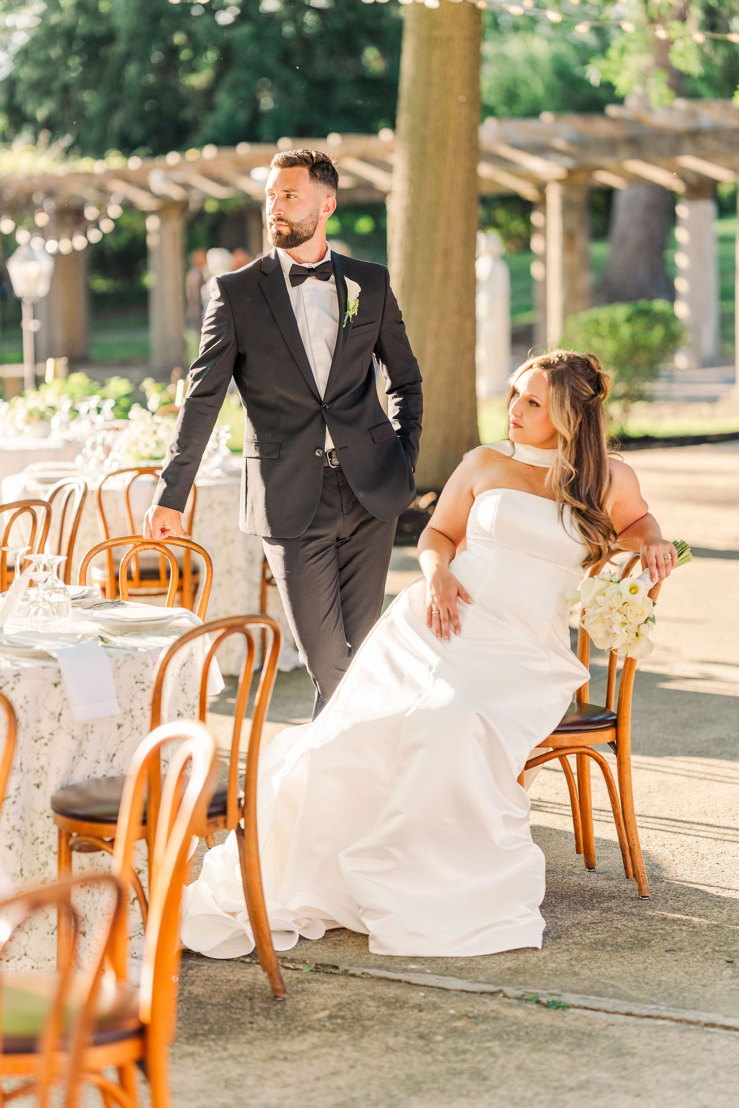Bride seated at outdoor reception table while groom in black tuxedo stands beside her in garden venue.