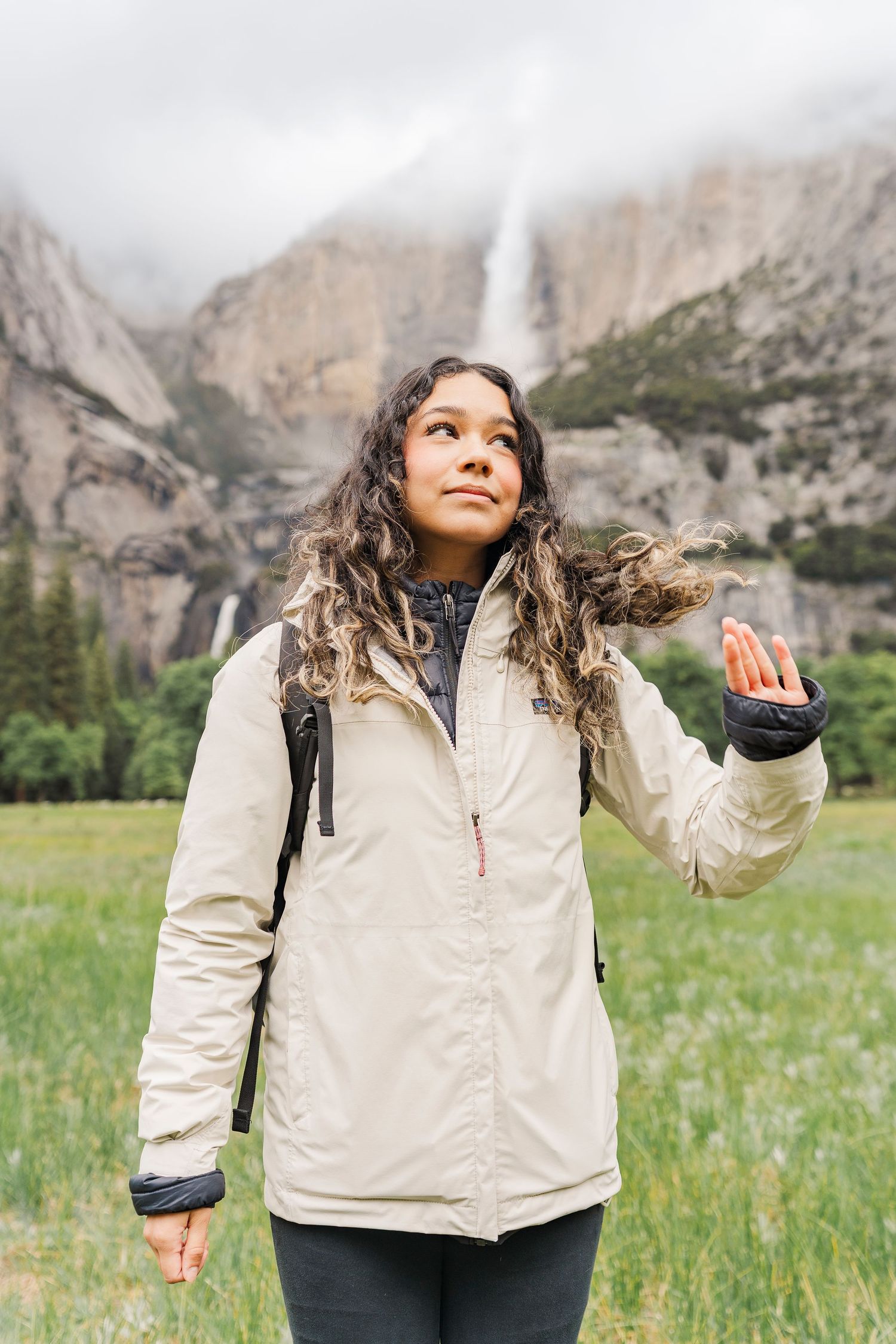 A young woman in a beige jacket looks up while hiking in Yosemite with a waterfall and cliffs behind her.