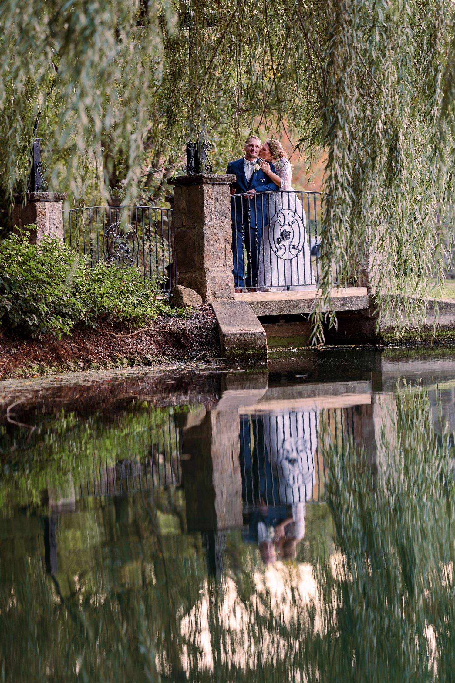 Couple in wedding attire standing on a stone balcony over a reflective pond beneath weeping willow trees at Gervasi Vineyard wedding venue.