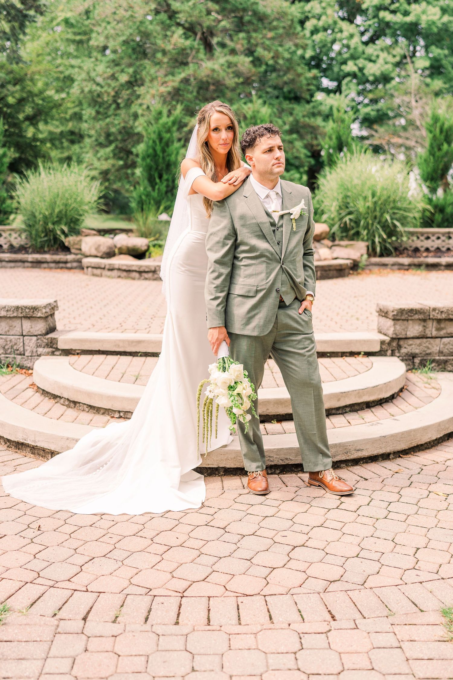 Bride in sleek white gown with long veil and groom in sage suit posing on brick courtyard steps.