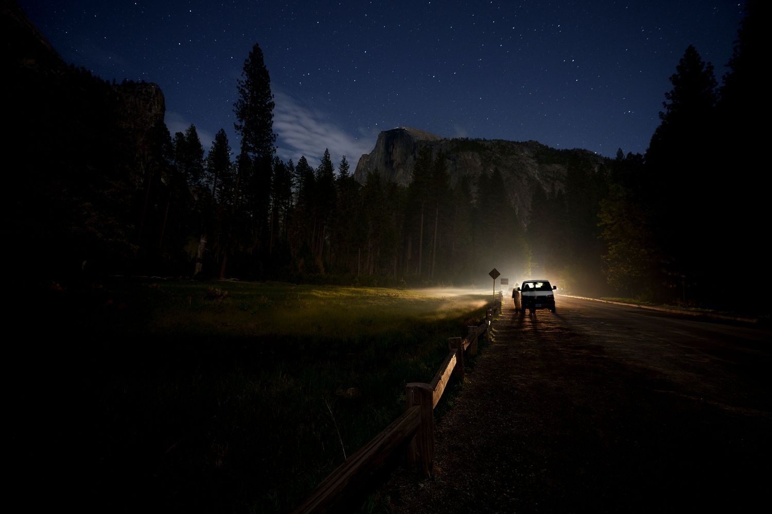 A car with bright headlights drives along a dark mountain road at night, surrounded by tall trees and a starry sky.
