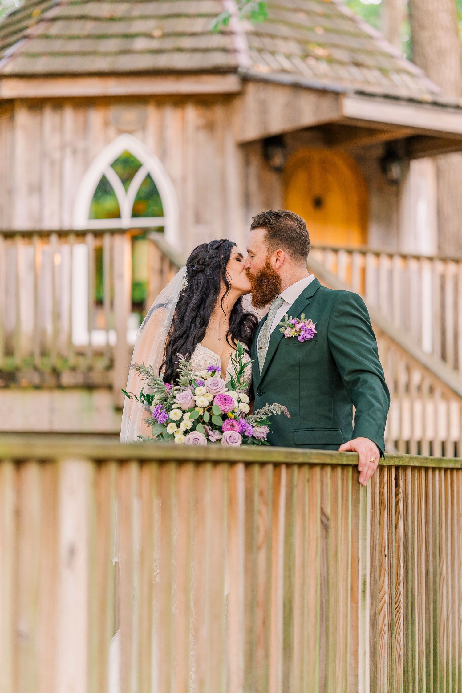 A bride and groom share a kiss on a wooden deck in front of a charming treehouse at their outdoor wedding at the Mohicans Treehouse Resort