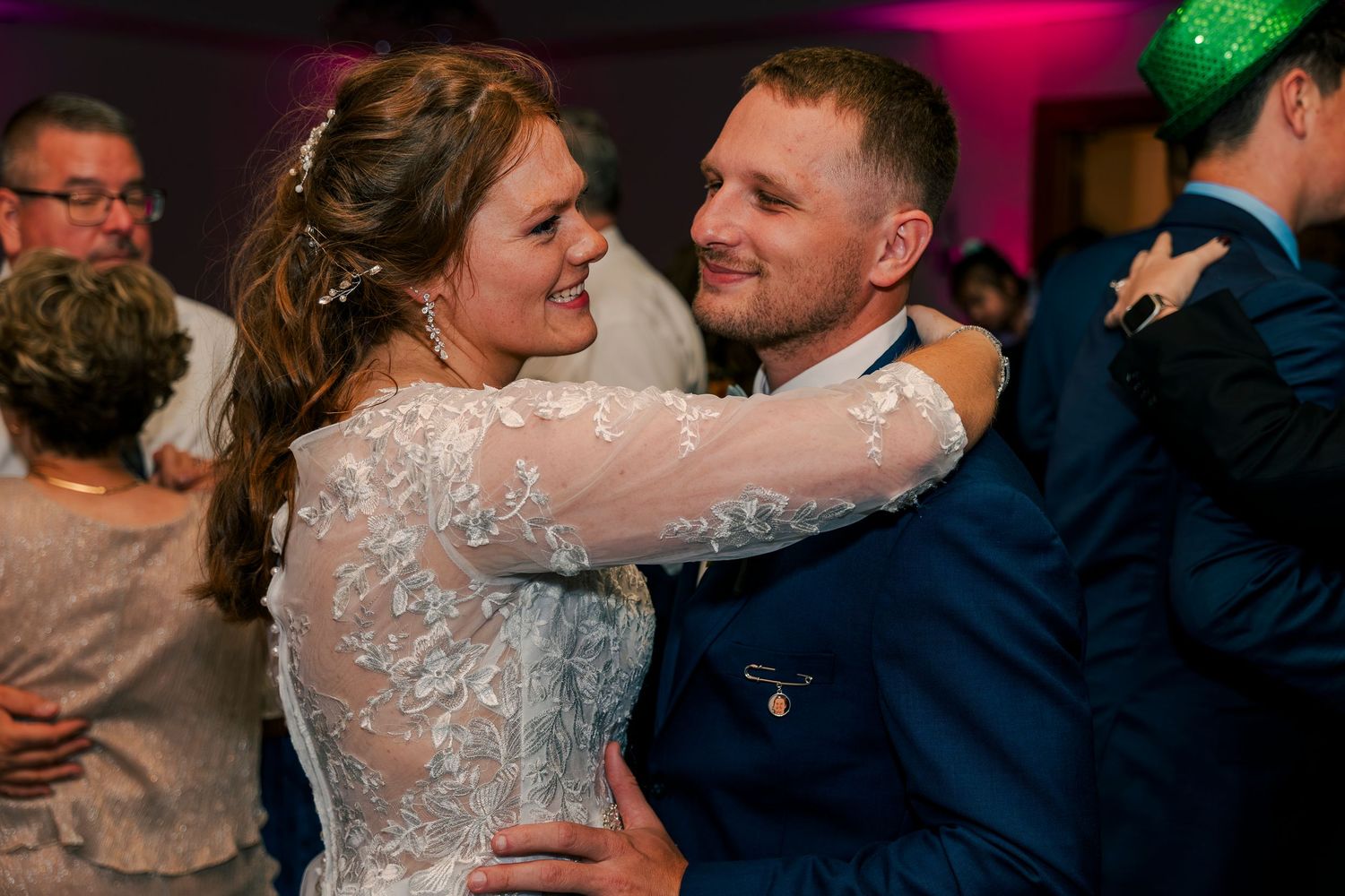 Bride and groom dance joyfully together surrounded by guests under colorful lights at wedding reception.