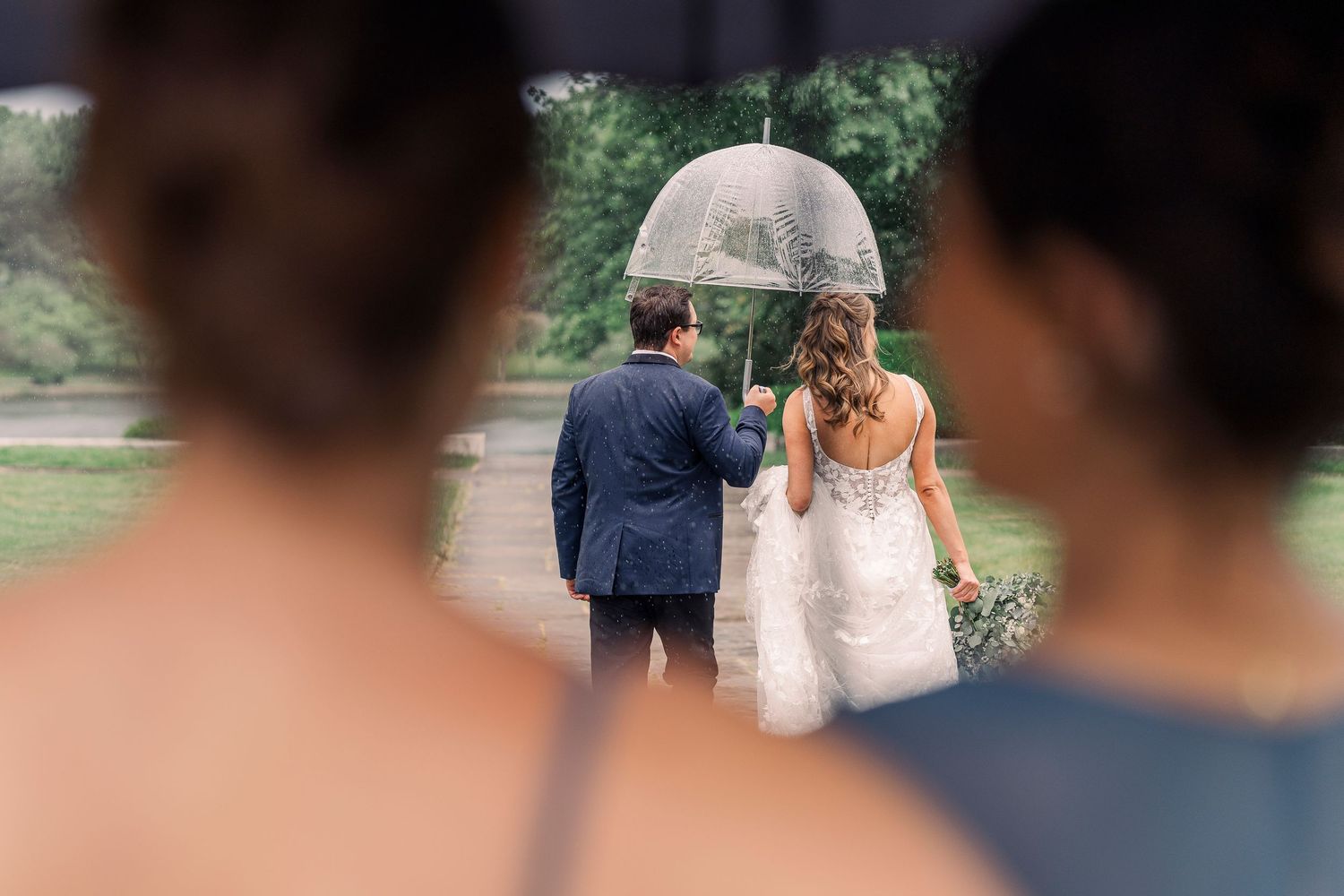 Bride and groom walking under a clear umbrella in the rain, viewed from behind by bridesmaids.