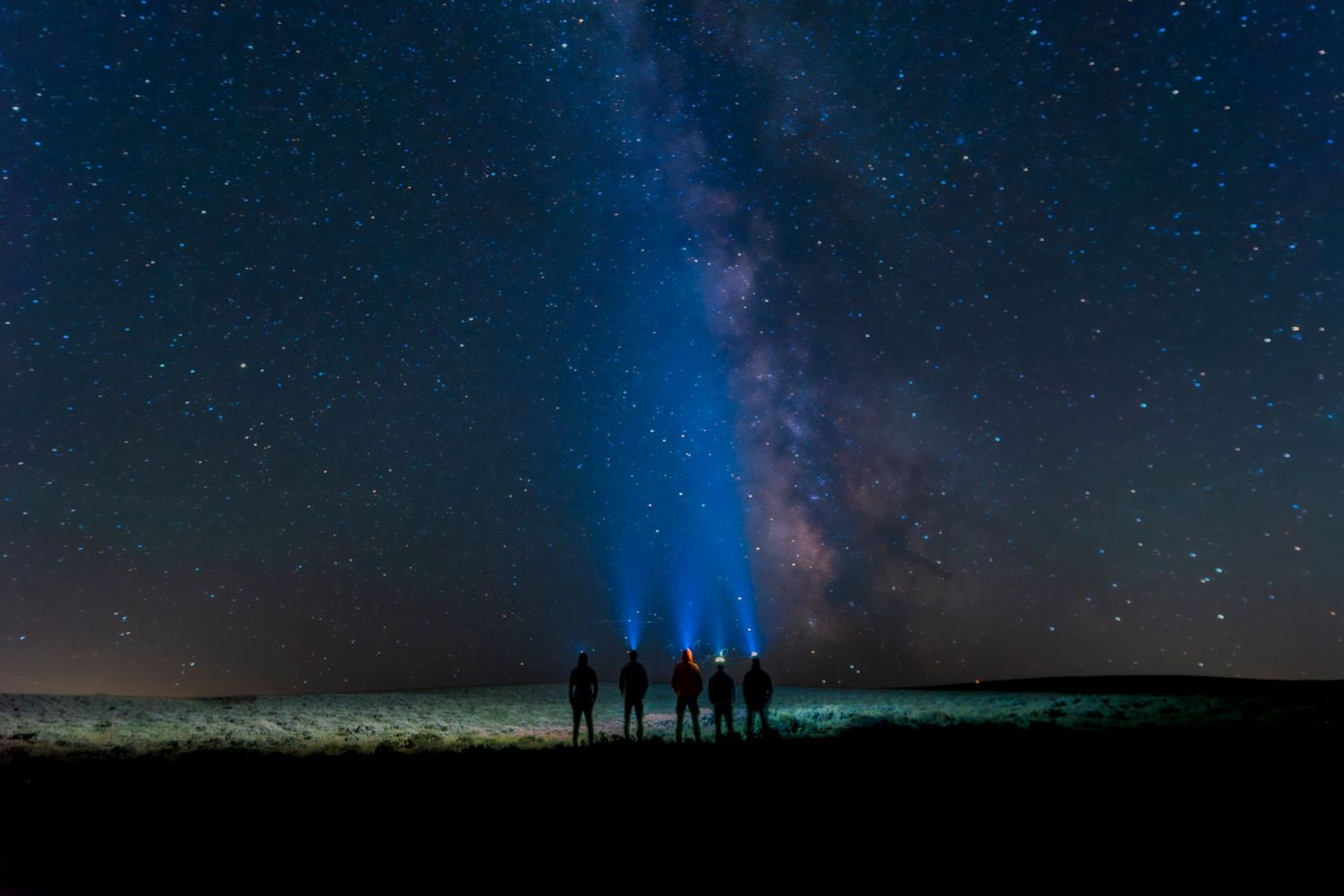 Silhouettes of five people shining headlamps toward the Milky Way galaxy on a clear starry night sky.