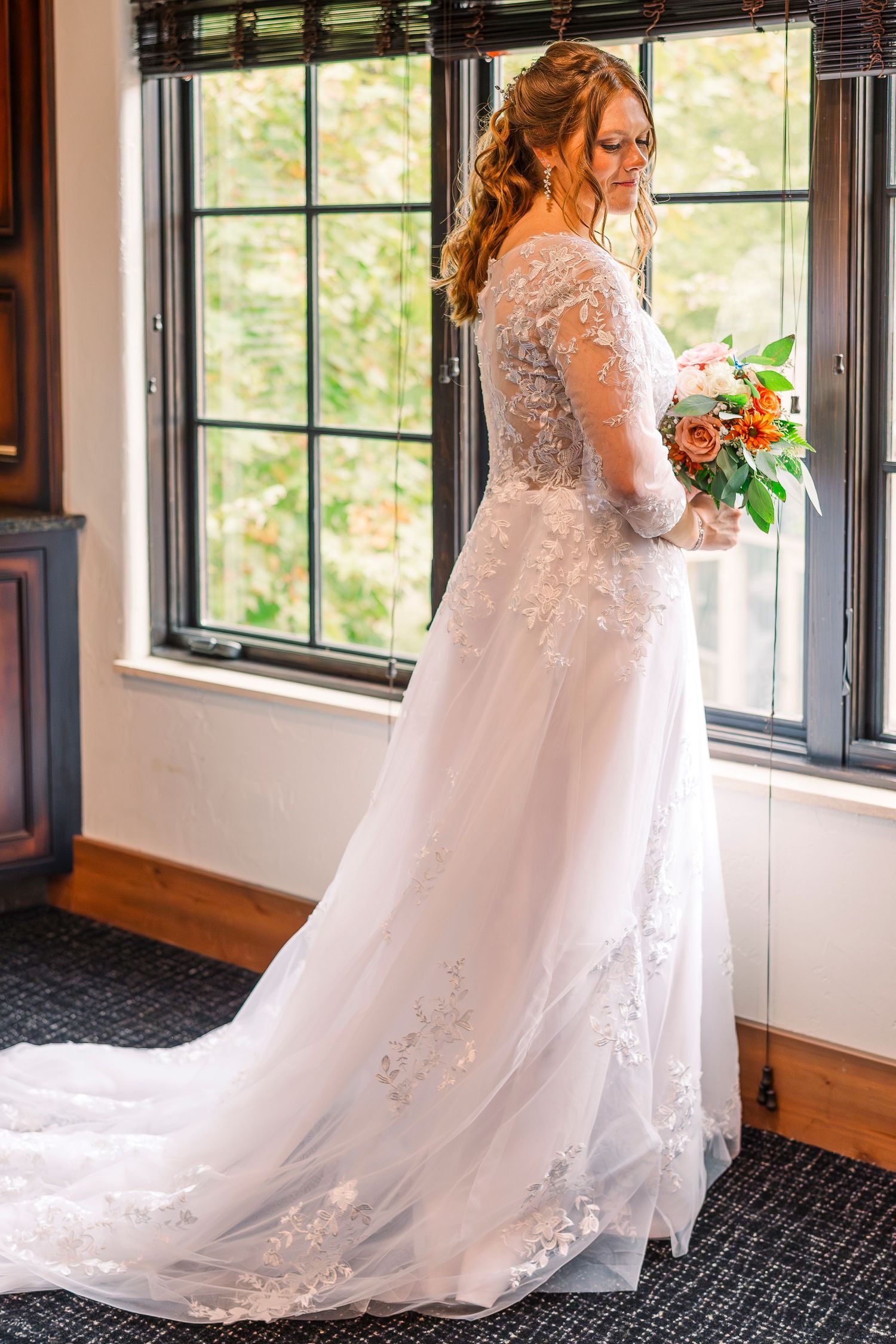 Elegant bride in lace wedding gown holding bouquet standing by large window.