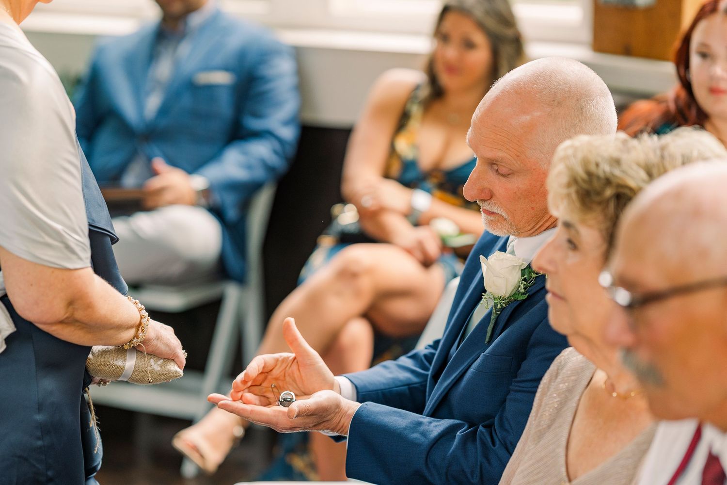 Wedding guest in navy suit holding ring box during indoor wedding ceremony.