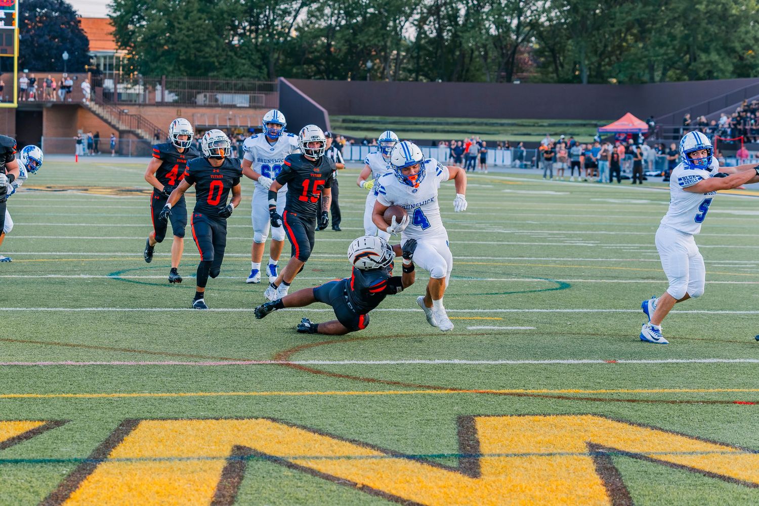 Brunswick High school football players in blue uniforms evade tacklers in red during an evening game on a yellow-marked field.