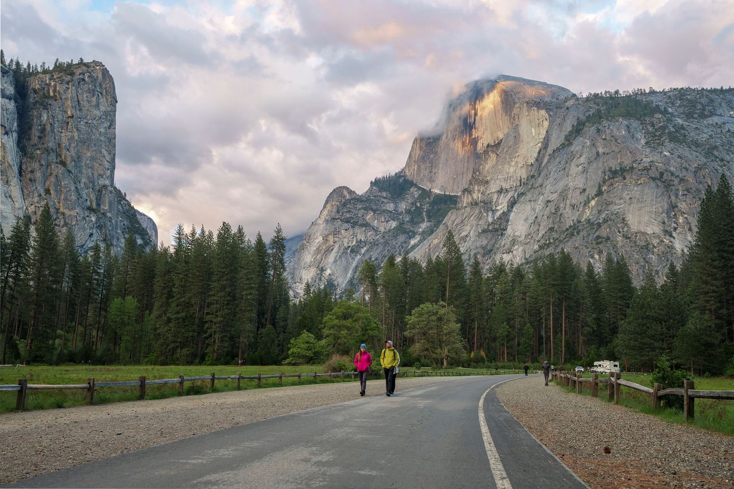 Two hikers walk along a scenic road in Yosemite Valley with Half Dome and granite cliffs under dramatic cloudy skies.