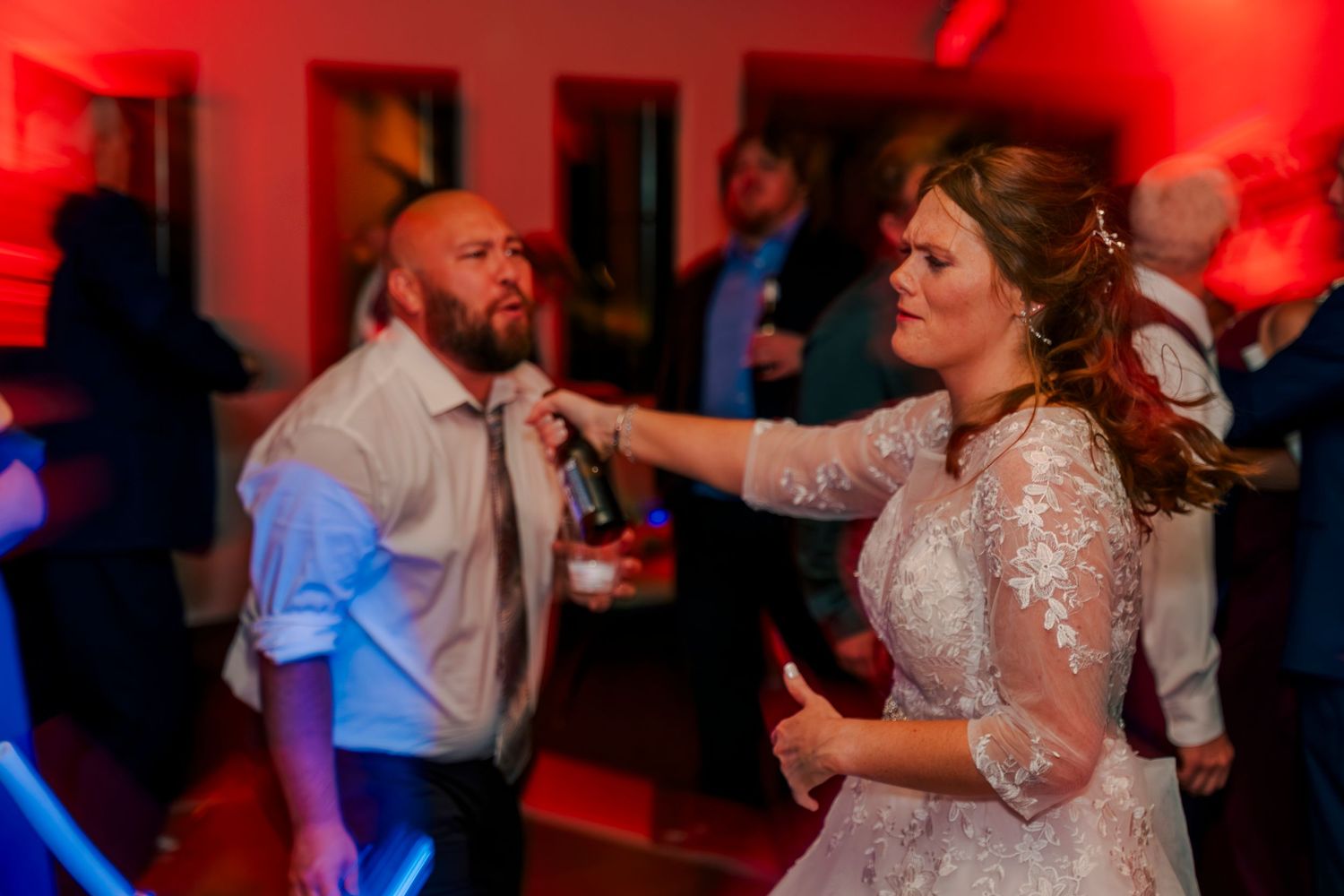 Bride in lace gown playfully holds microphone while dancing with guest under red lights at reception.