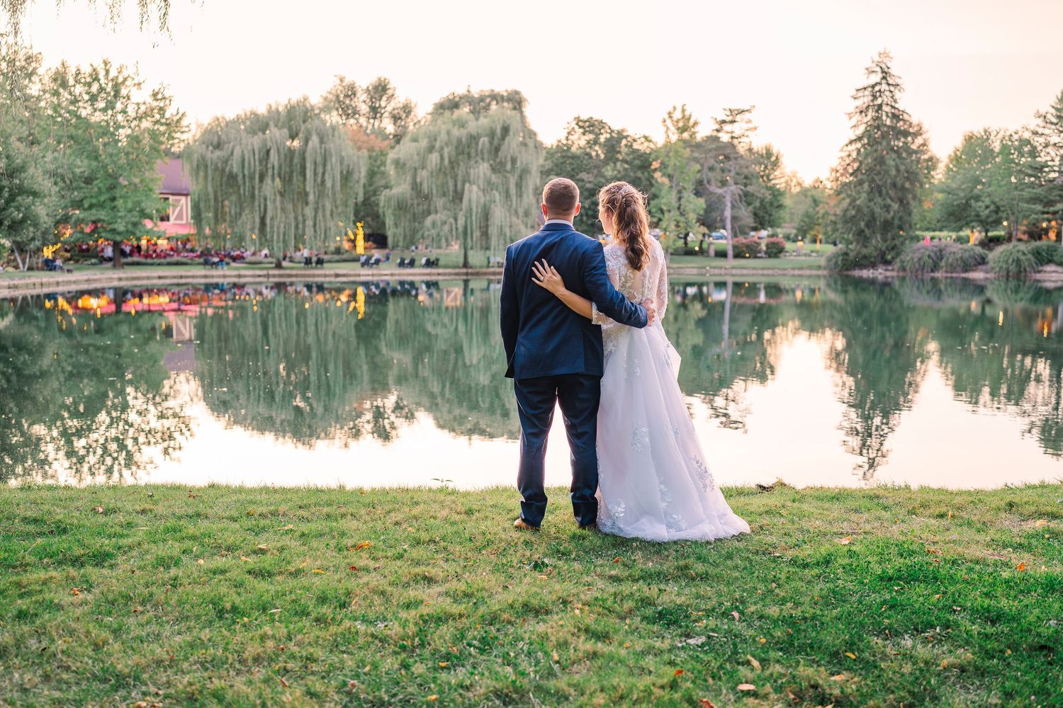 Bride and groom embracing while gazing at a tranquil sunset pond surrounded by willow trees at Gervasi Vineyard wedding venue.