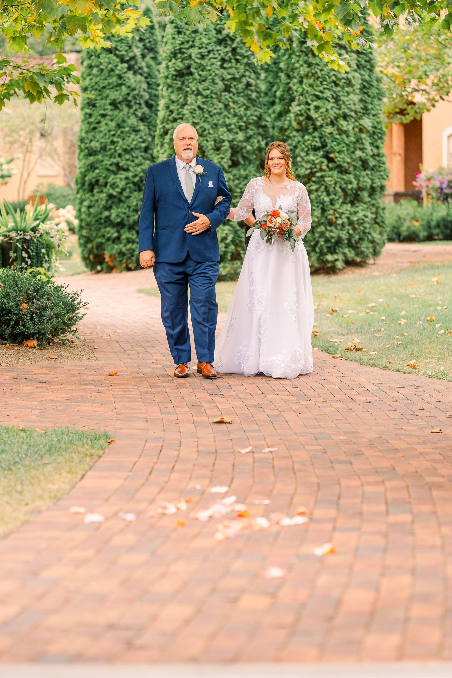 Bride holding colorful bouquet walks with older man in navy suit down brick aisle lined with hedges.