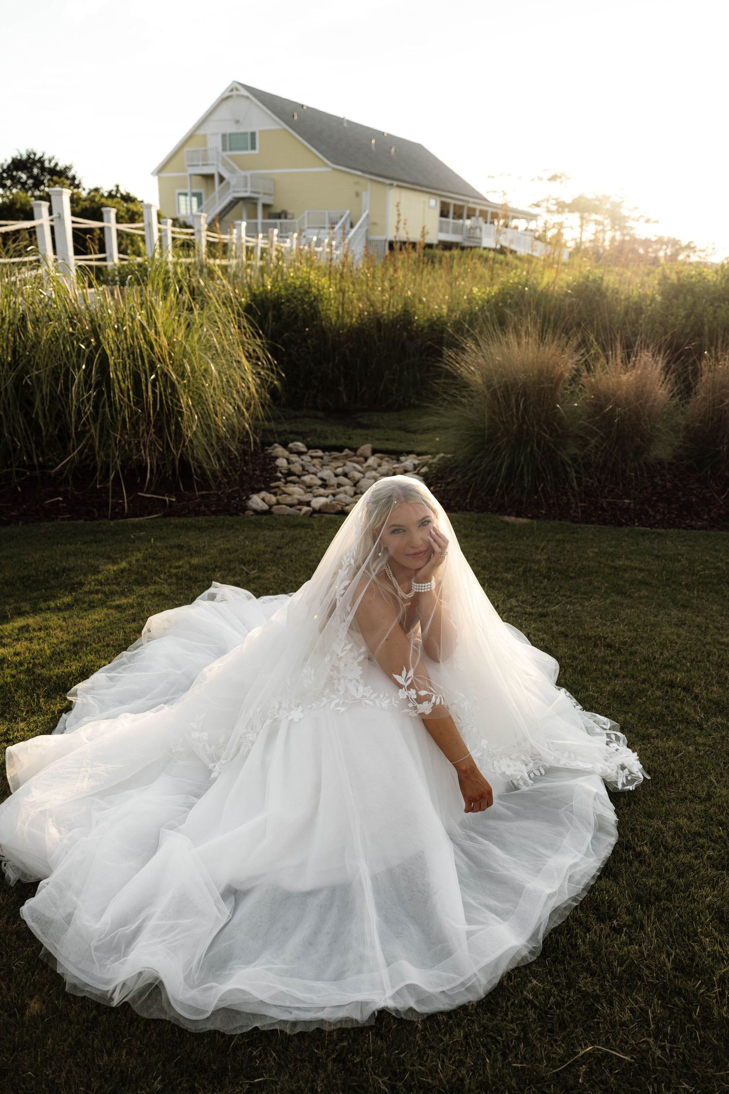 Bride in a flowing white ballgown and veil sits on green grass outside a coastal house at golden hour.