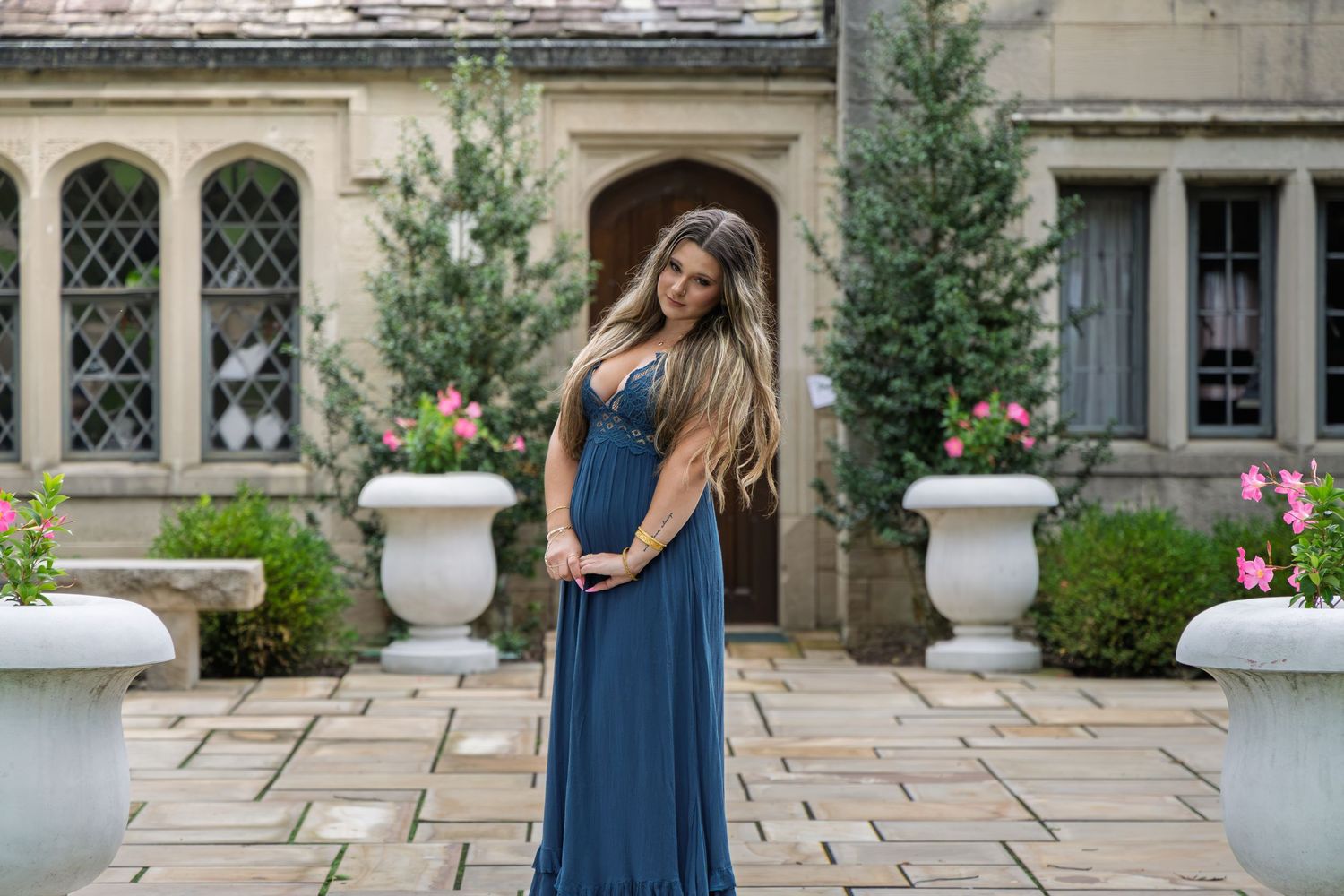 Young woman in flowing blue gown stands on stone patio of a grand manor house.