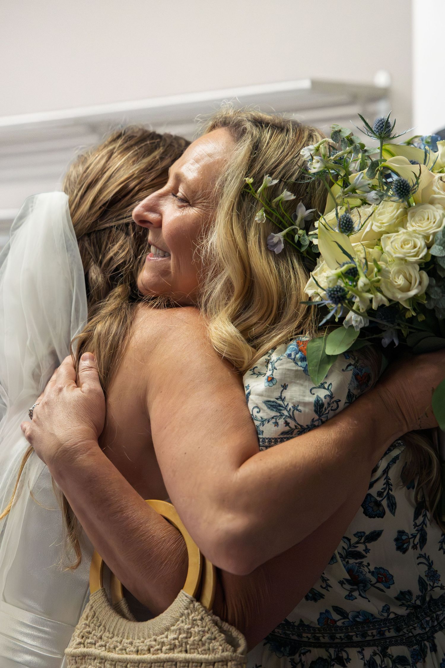 Bride and older woman share emotional hug at wedding, bouquet of white roses and blue flowers visible.