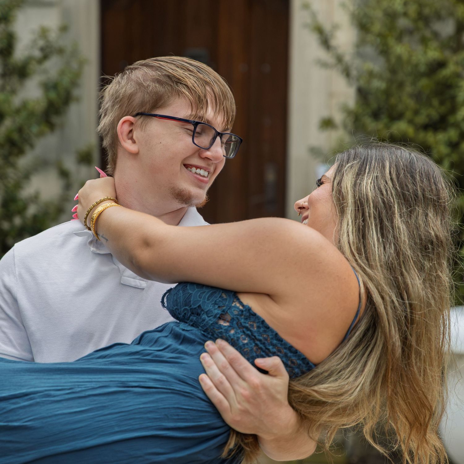 A smiling young man in glasses lifts a laughing blonde woman in a teal dress outdoors.
