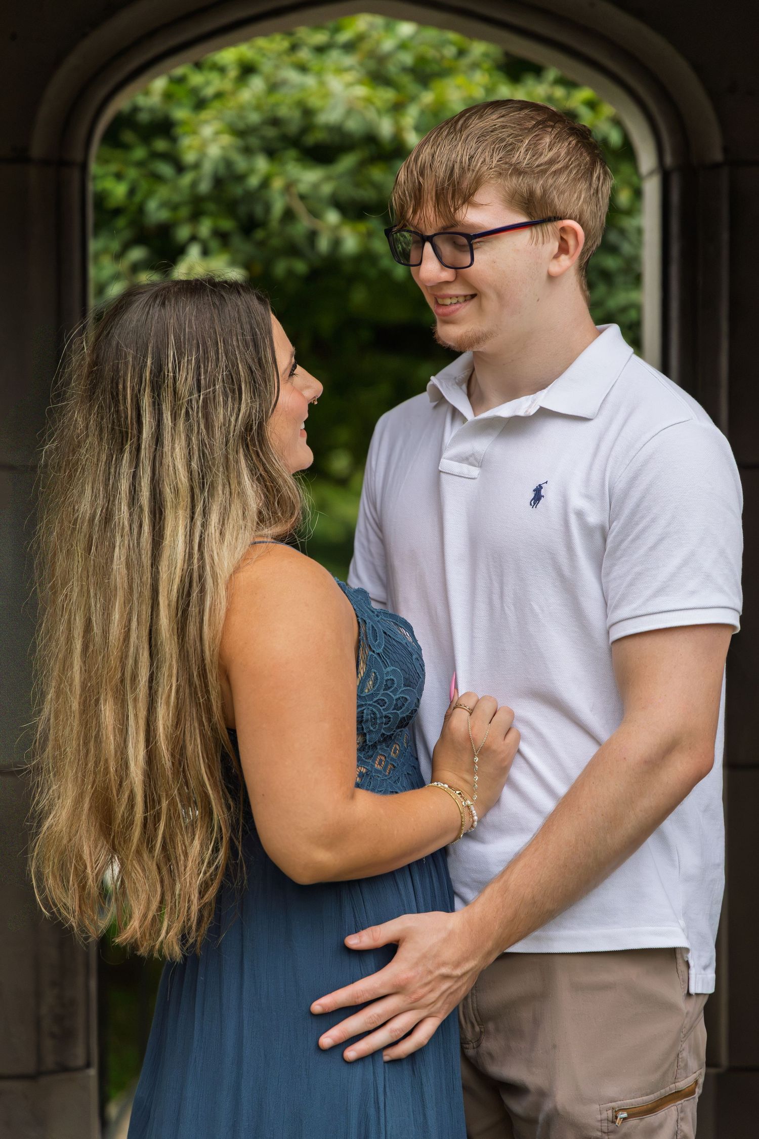 A young couple in a romantic embrace, smiling at each other, outdoors near a stone arch with lush greenery.