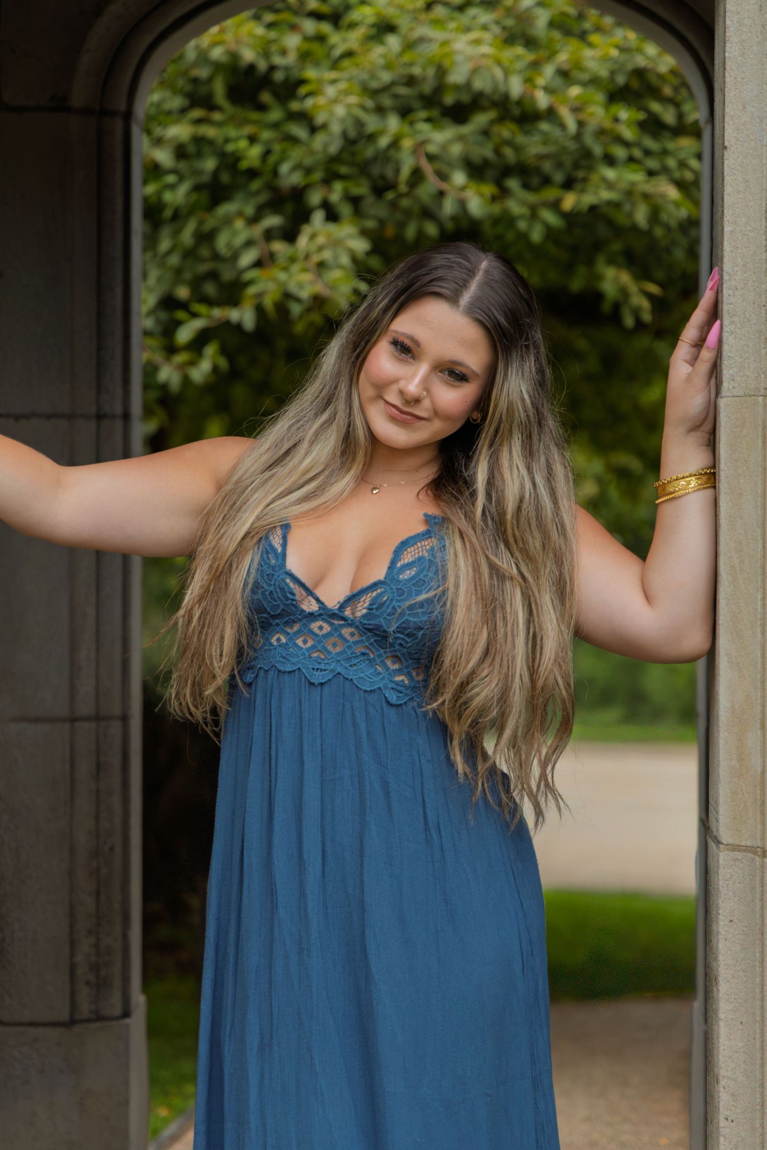 Young woman with long blonde hair poses in a blue lace maxi dress under a stone archway with lush greenery.