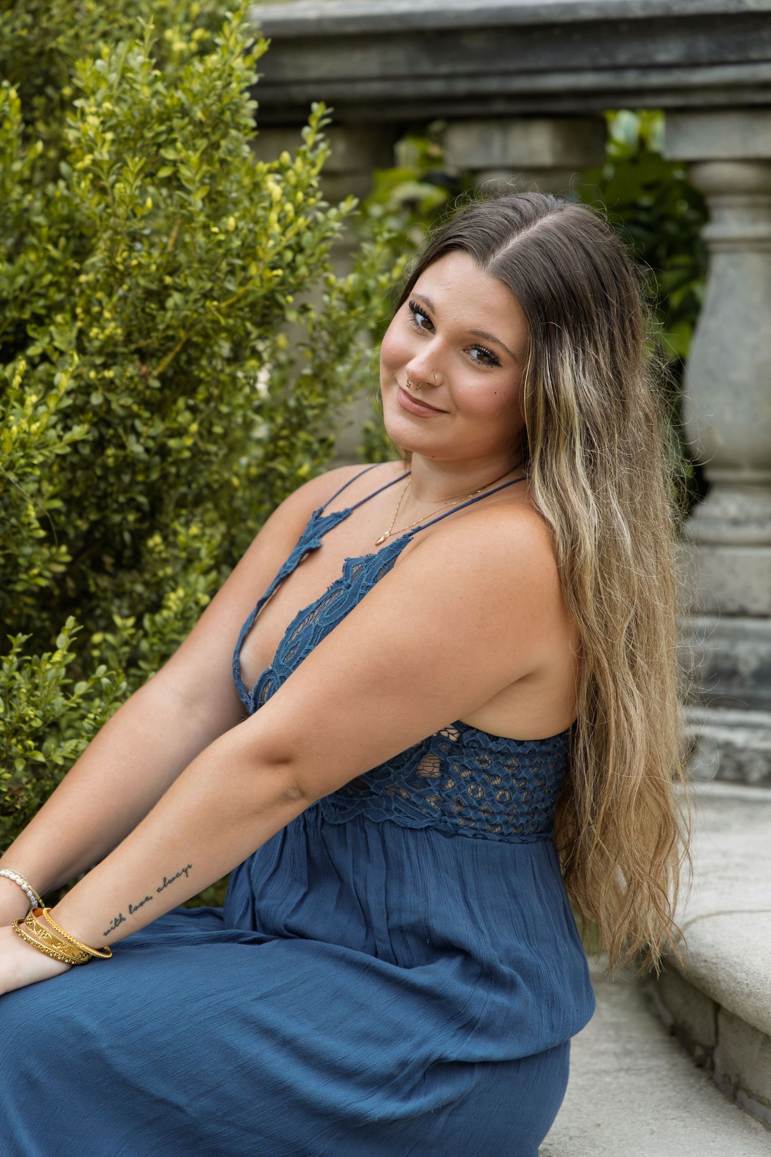 Young woman with long hair sits outdoors in a blue lace dress, smiling over her shoulder near stone columns and green shrubs.
